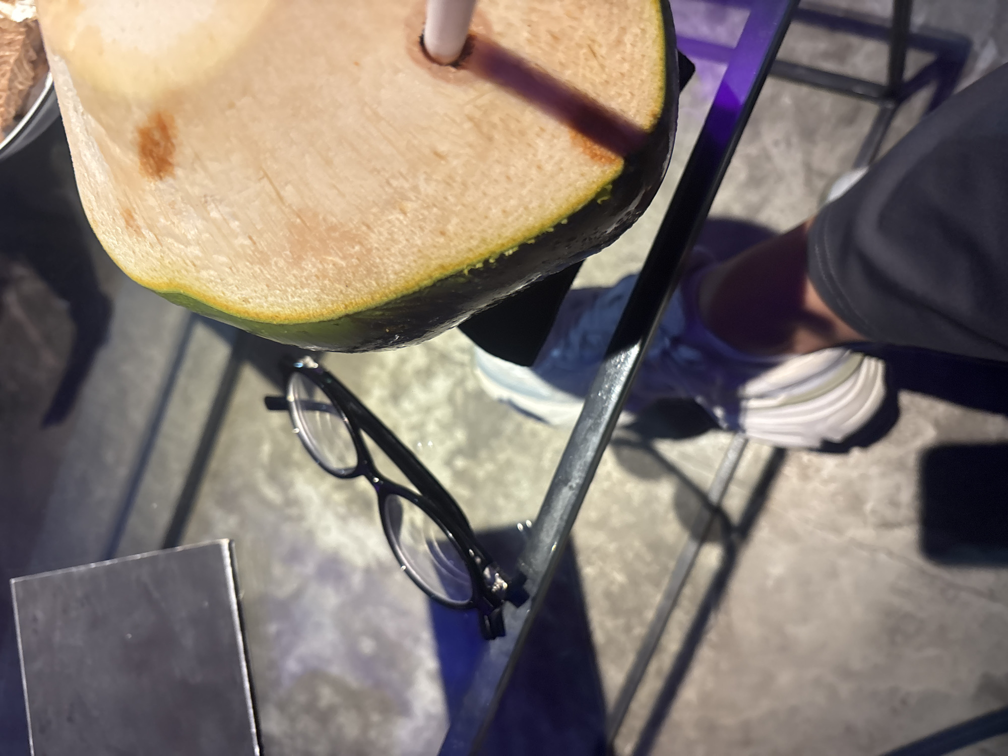 A close-up, overhead shot shows a person's legs and feet wearing white sneakers and dark pants, positioned on a grey floor. They are seated at a glass-top table with a metal frame, on which rests a pair of black-rimmed glasses. To the right, half of a coconut with a straw is visible, with a brown stain on its flesh. A black metal object is on the left side of the table. The lighting appears to be dim, with purple accents from under the table. The scene is indoors, in an unidentifiable urban setting.
