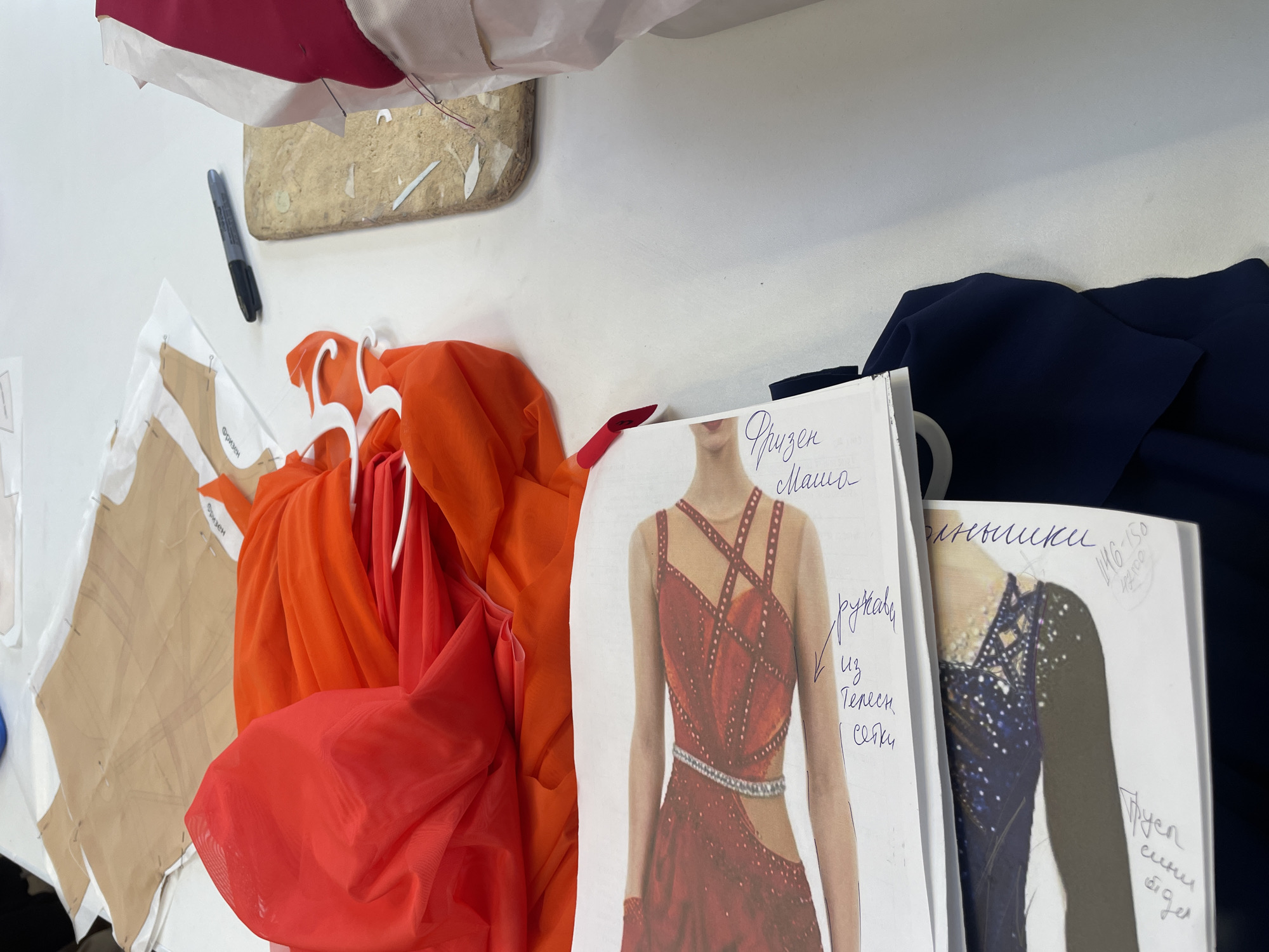 A flat lay view of a sewing workspace on a white surface shows fabric patterns, red and orange sheer fabric draped on hangers, a marker pen, and sketches of figure skating costumes. The background is out of focus. The image is taken from a slightly elevated angle, looking down at the table. No people are visible. The environment appears to be an indoor workshop, possibly a studio or a room in a residential building. The context of Sinyavino, Russia, is not visually evident from the image itself.