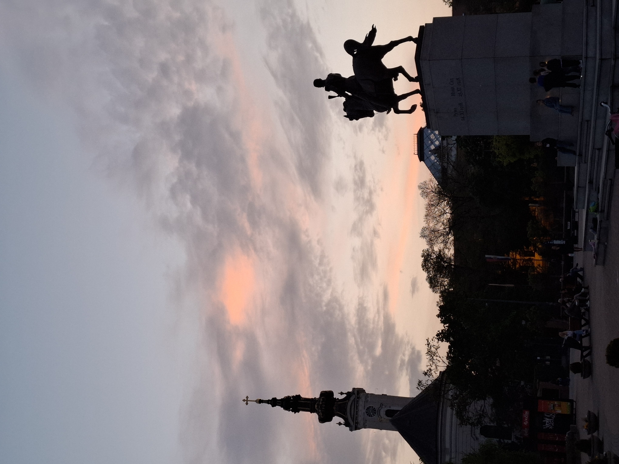 The image, taken at dusk, showcases the iconic landmarks of Novi Sad, Serbia.
Dominating the scene is the statue of Svetozar Miletić, silhouetted against the colorful sky. The statue is mounted on a stone pedestal with people gathered around.
In the foreground, the distinctive spire of the Name of Mary Church (also known as the Novi Sad Cathedral) pierces the sky. The surrounding area features trees and cityscape.
The overall atmosphere suggests a blend of history, culture, and urban life in Novi Sad.