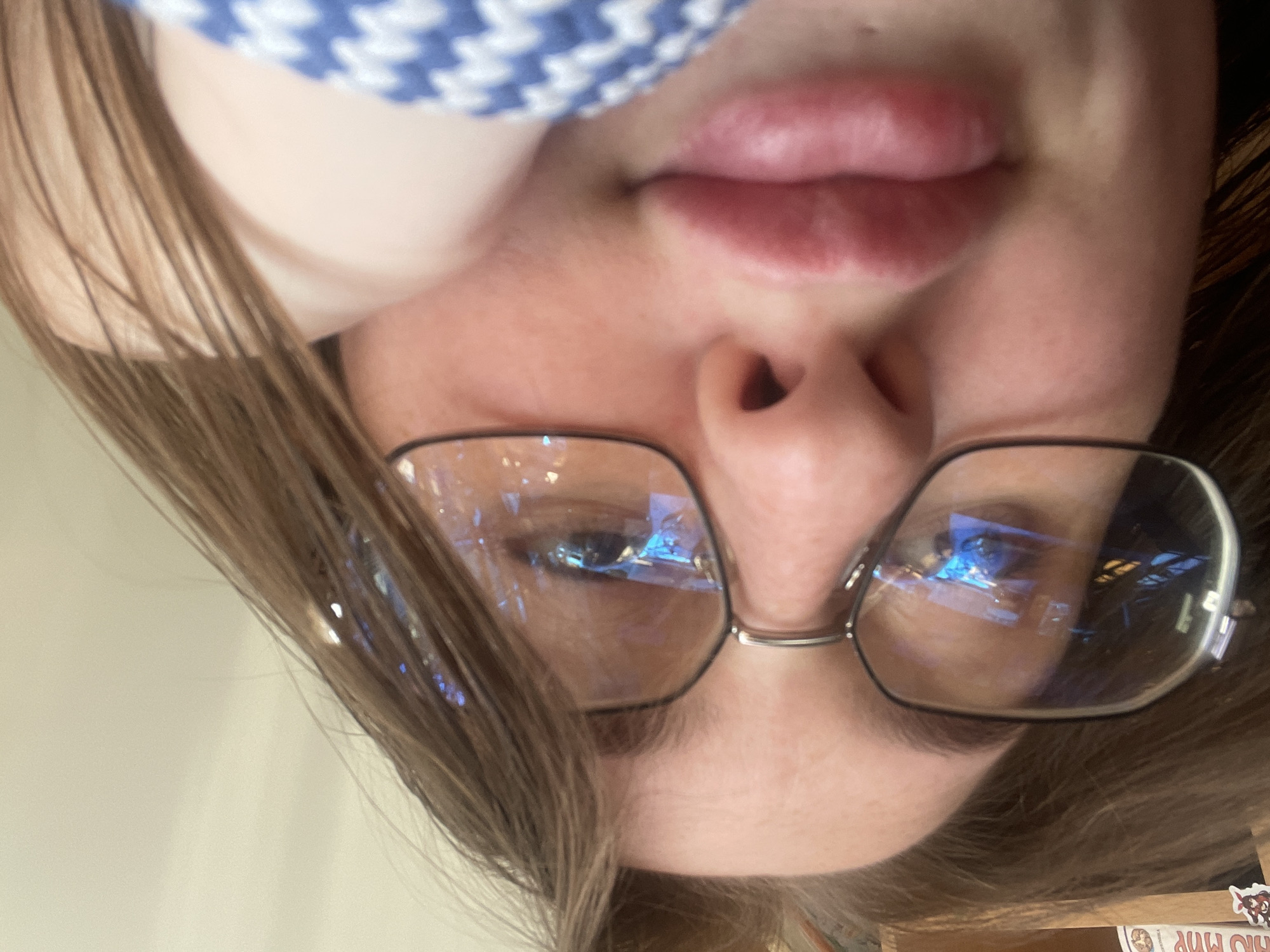 The image is a close-up, low-angle selfie of a person wearing glasses. The person's face is partially obscured by their hand and hair. Their lips are prominent and appear to be pursed or slightly parted. The background is indistinct, but the glasses reflect what appears to be a screen or monitor displaying bright blue and white light, possibly a computer screen. The environment is indoors, suggested by the soft lighting and the reflection in the glasses. There are no discernable elements within the image that would indicate a specific location within the city of Сырково, Russia.
