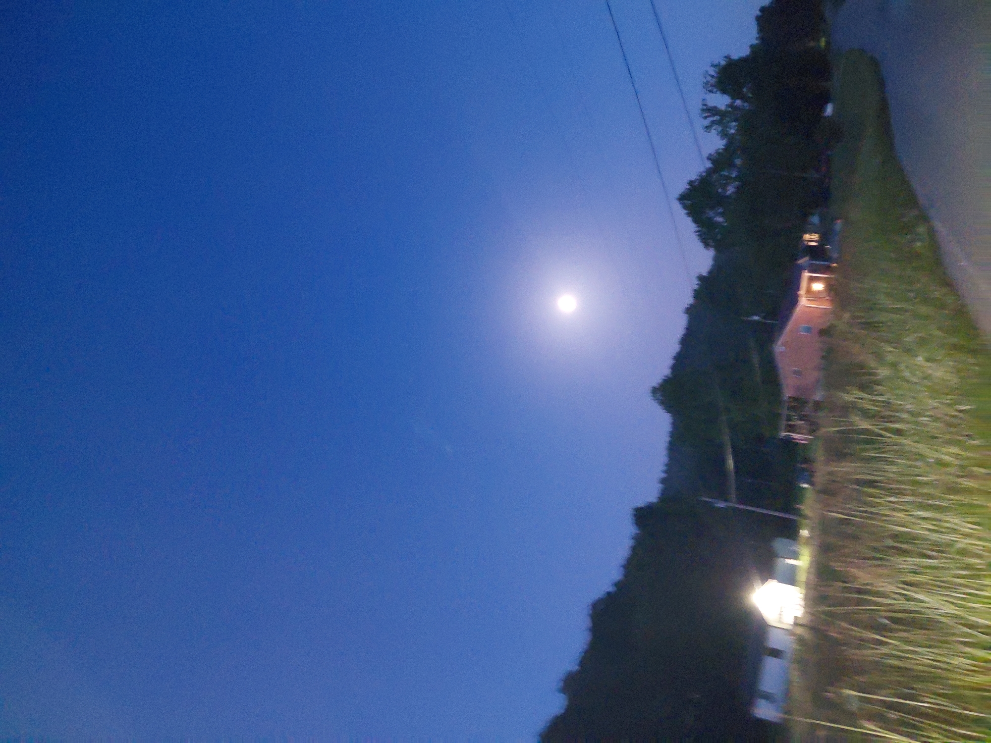 This media file captures a twilight scene in Berea, United States of America, likely during the evening. The sky is a deep, clear blue, transitioning from dusk to night. A bright, luminous full moon is prominently visible in the upper right quadrant of the frame, casting a soft glow.
The scene appears to be a rural or suburban setting, with a dense line of trees dominating the right side of the image, obscuring much of the background. The trees suggest a natural landscape, possibly a wooded area bordering a residential or developed area.
In the lower right, a paved surface, likely a road or driveway, is partially visible, edged by tall, dry grass or reeds that are illuminated by artificial light. A streetlamp, emitting a warm, bright light, stands near the road. Further back, a building with illuminated windows is visible among the trees. The architecture suggests a house or a small structure.
The overall mood is tranquil and serene, enhanced by the soft moonlight and the quietude implied by the twilight setting. The image has a slightly tilted perspective, adding a dynamic quality to the composition. No people are visible in the frame, and there is no visible text. The weather appears clear and calm.