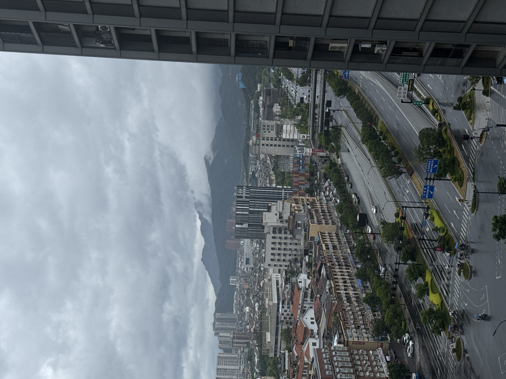 The image is an aerial view of a rainy day in Yiwu, China, likely taken from a high-rise building. In the foreground, a modern, dark-colored high-rise building dominates the right side of the frame. Its facade features repetitive balconies and windows. Below, a multi-lane road intersection is wet from the rain, with cars and motorcycles navigating through it. Pedestrians with umbrellas can be seen crossing the street. Further into the cityscape, a dense collection of buildings, ranging from multi-story residential blocks to commercial structures with varied architectural styles and roof colors, stretches across the middle ground. Some buildings have awnings or balconies. A prominent building with a striped facade stands out. Beyond the urban sprawl, a range of green, mist-shrouded mountains forms the backdrop, indicating the city's proximity to natural landscapes. The sky is overcast with heavy, gray clouds. Traffic lights and road signs are visible throughout the scene. The overall impression is of a bustling city enduring inclement weather.