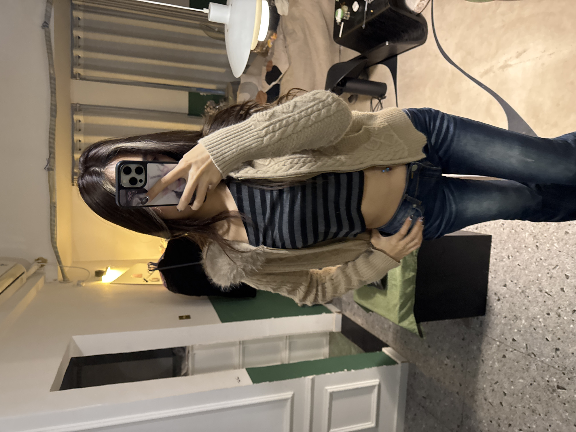 The image portrays an indoor scene located in Liangjiang, China, featuring an individual taking a mirror selfie. The person, positioned centrally, holds a black-cased smartphone to obscure their face, with the right hand visible gripping the device. The left hand rests on the hip. The individual wears a beige cable-knit cardigan with a fur-like collar, a black and grey horizontal striped crop top exposing the midriff and a navel piercing, and dark blue jeans. The visible hair is long and dark with lighter streaks framing the face.
The flooring in the foreground consists of light-colored terrazzo or speckled concrete. To the immediate left, a white wall section features decorative molding and a lower dark green panel, alongside an open doorway leading to an unseen area. An overhead air conditioning unit is mounted on the wall above this section. In the background, a bed with light-colored, disheveled bedding is visible. Adjacent to the bed, a dark-colored nightstand holds several small objects, and a white-shaded pendant lamp hangs above the bed area. Striped curtains cover a window or opening further in the background. A dark garment hangs on a hook against a green wall section. A dark, rectangular object covered with a green cloth rests in the mid-ground, and another dark object is on the floor to the right. Warm artificial lighting illuminates the room from multiple sources.