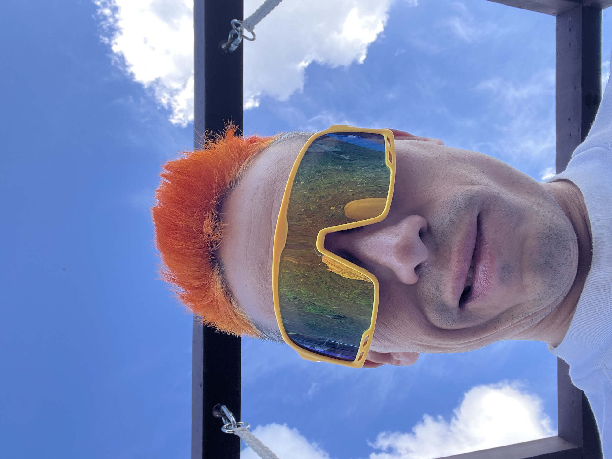 A low-angle, close-up shot captures a person wearing large yellow sunglasses that reflect a landscape. The person has orange hair and is wearing a white shirt. The background is a clear blue sky with scattered white clouds, along with parts of a dark-colored structure with ropes attached. The location is potentially outdoors in the city of Chaikovsky, Russia.