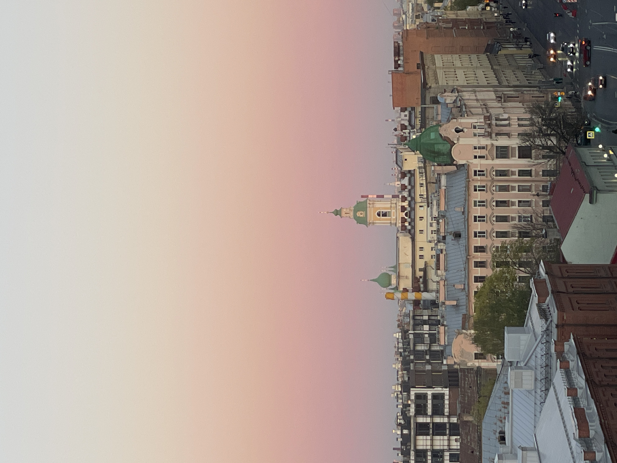 The image shows a view of Saint Petersburg, Russia, at dusk or dawn. The sky is a gradient of pale pink and light gray, suggesting a sunrise or sunset. The city skyline is dominated by traditional architecture with several buildings and rooftop views. Two prominent buildings have green domes. On the right, one street has vehicular traffic, with cars and street lamps visible. The overall impression is that the picture was taken from an elevated position, possibly a rooftop or high window, providing a wide-angle view of the city.