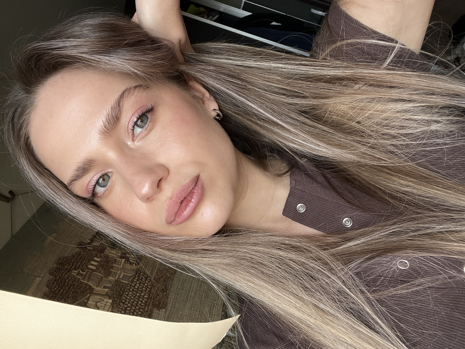 The image displays a head and shoulders self-portrait of a female subject, stated to be located in Saint Petersburg, Russia, within an indoor environment. The woman has fair skin, light green eyes, prominent eyebrows, and full lips colored with a light pink product. Her long hair, a mix of blonde highlights and darker tones, cascades over her shoulders and chest. She wears a dark brown, ribbed top with silver snap buttons along the front placket. A small silver hoop earring is visible in her right ear. Her right hand is partially visible at the upper right, resting on or near the back of her head. In the background to the left, a light beige fabric element (possibly a curtain or blind) is partially drawn, revealing a tapestry or wall hanging behind it that depicts a landscape scene with buildings and trees in earthy tones. The background to the right is darker and indistinct, with faint shapes suggesting interior furnishings.