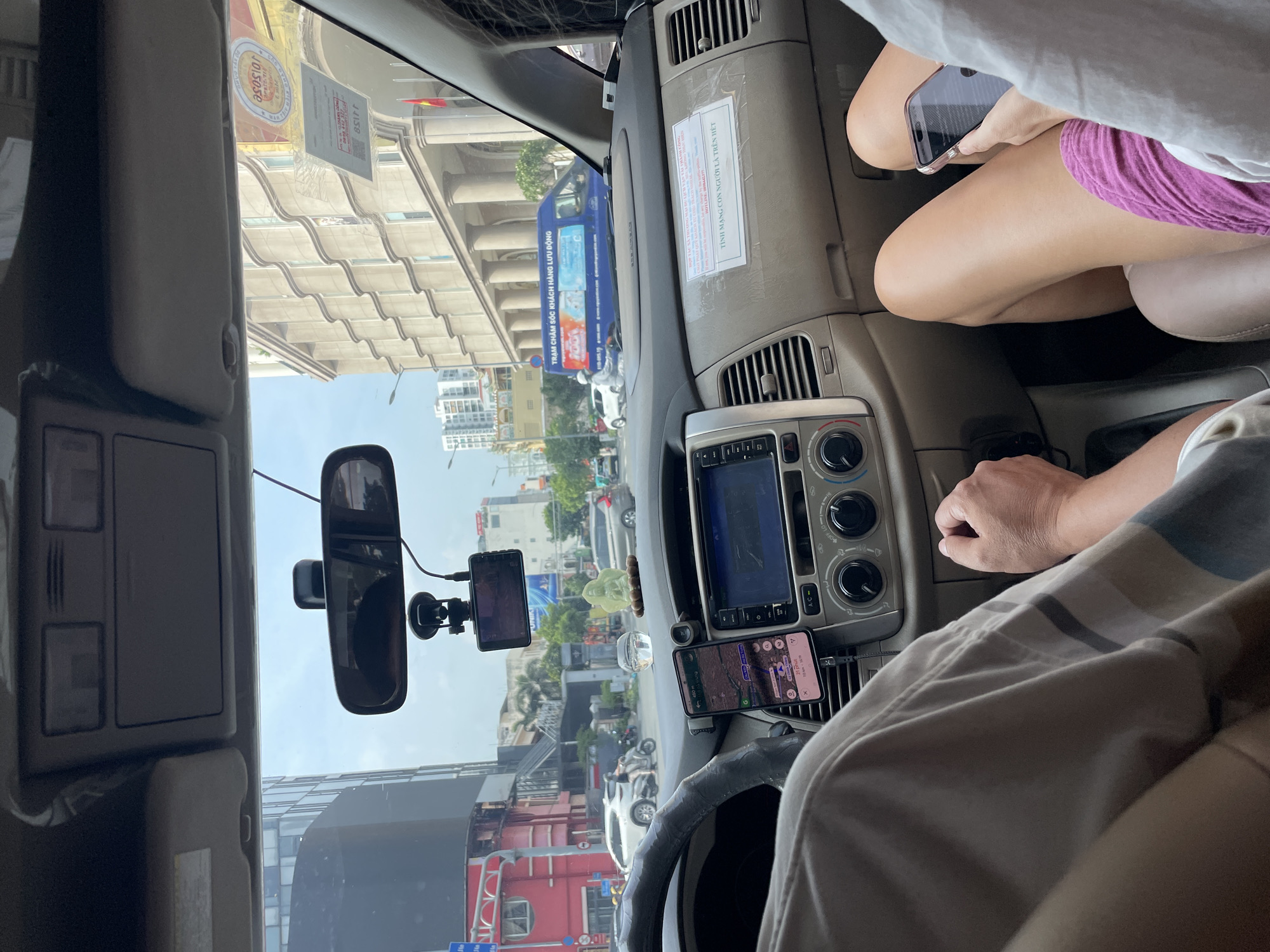 The image shows the interior of a car, viewed from the passenger's perspective, looking towards the dashboard and out the windshield. The car appears to be a Toyota Innova, common for taxi services in Vietnam. In the foreground, the driver's left arm and shoulder are visible, resting on the steering wheel, along with the passenger's legs. A smartphone is mounted on the dashboard, displaying a navigation app, likely Google Maps, with the route indicating a destination in Thủ Đức, Vietnam. The dashboard features a central infotainment screen, climate controls, and air vents.
Outside the car, a bustling city street is visible under an overcast sky. Modern buildings with classical architectural elements line the street, suggesting an urban environment. Several vehicles, including cars, motorcycles, and a blue commercial van, are present on the road, indicative of traffic. A traffic light is visible on the left side of the frame. The overall scene depicts a typical daily commute or travel scenario within the city of Thủ Đức.