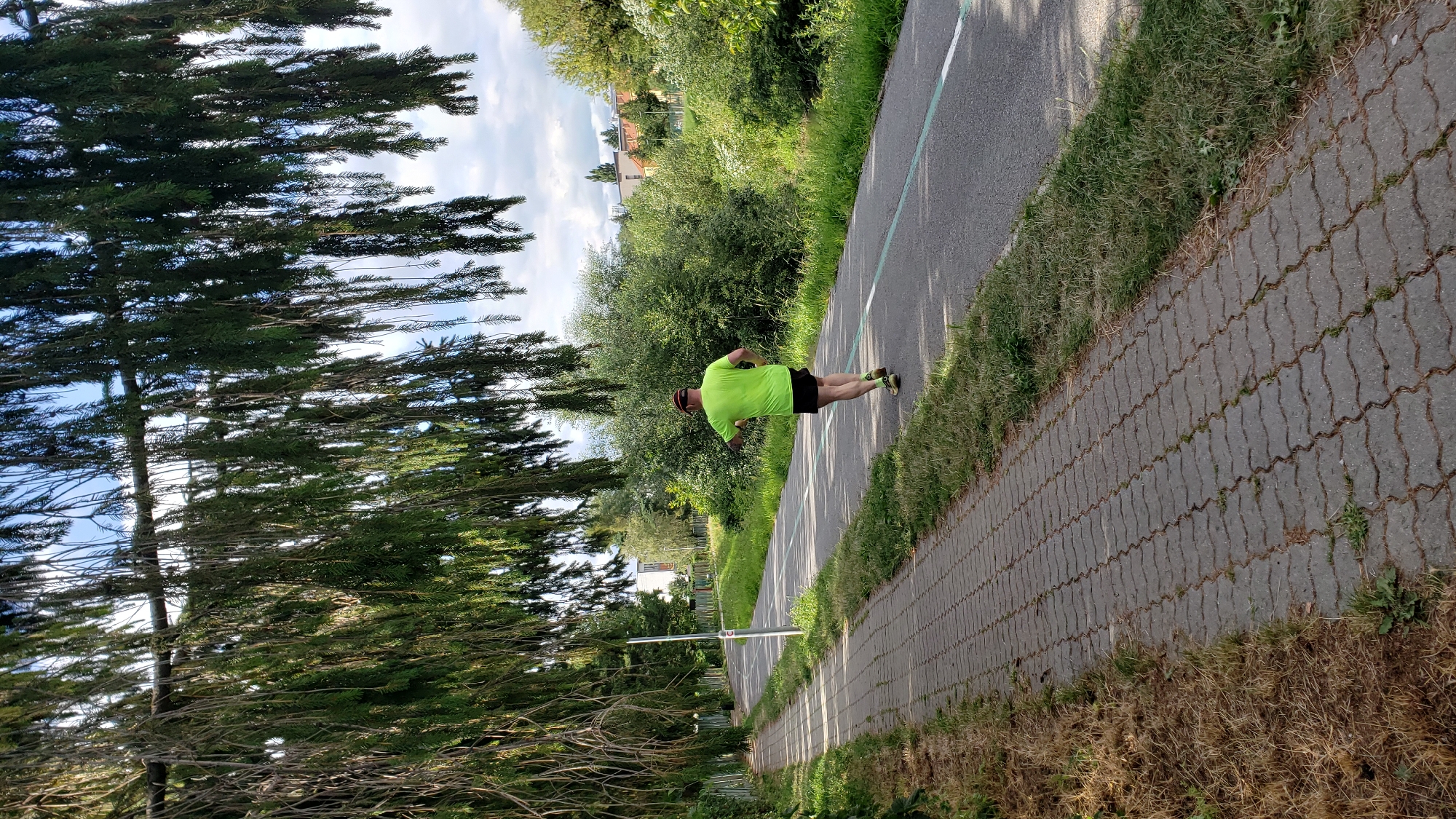 A man in a bright yellow t-shirt and black shorts runs on a paved road with a green stripe, which appears to be a bike path or dedicated running lane, in Prešov, Slovakia. He is wearing a red cap and athletic shoes. The path is to the right of a paved pedestrian walkway lined with dry grass. To the left of the pedestrian walkway is a metal fence and lush green trees. The background consists of dense green foliage, buildings in the distance, and a cloudy sky. The image is taken from behind the runner, looking forward along the path.