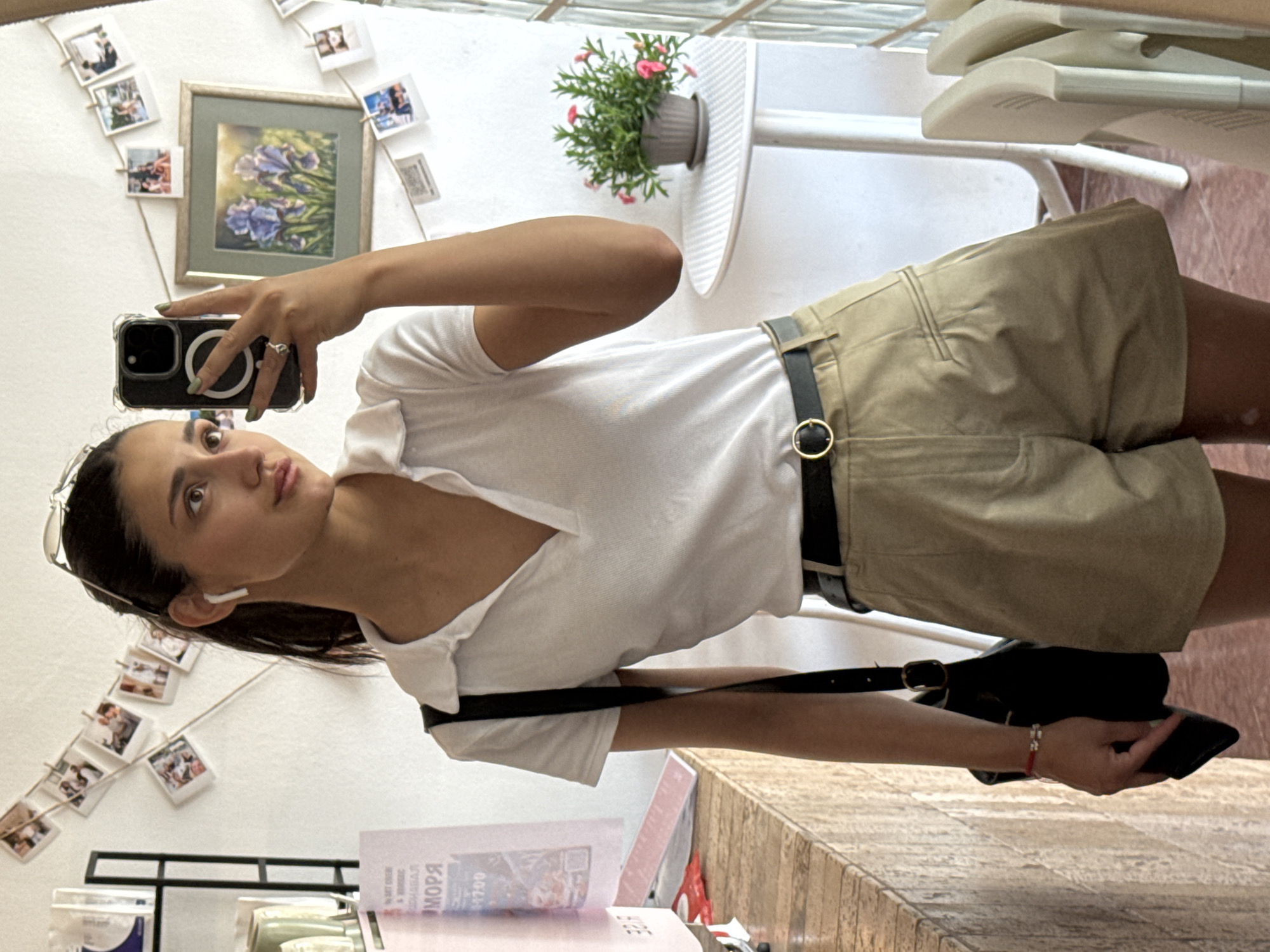 A woman is taking a selfie in what appears to be an indoor setting, possibly a café or restaurant. The background includes framed pictures on the wall and a table with a potted plant. The woman is wearing a white polo shirt and khaki shorts with a black belt, and she has a black purse or bag slung over her shoulder. She holds her smartphone up to take the photo, and her expression is neutral. The counter space in the front of the image is marbled. The image was taken in Bar, Montenegro.