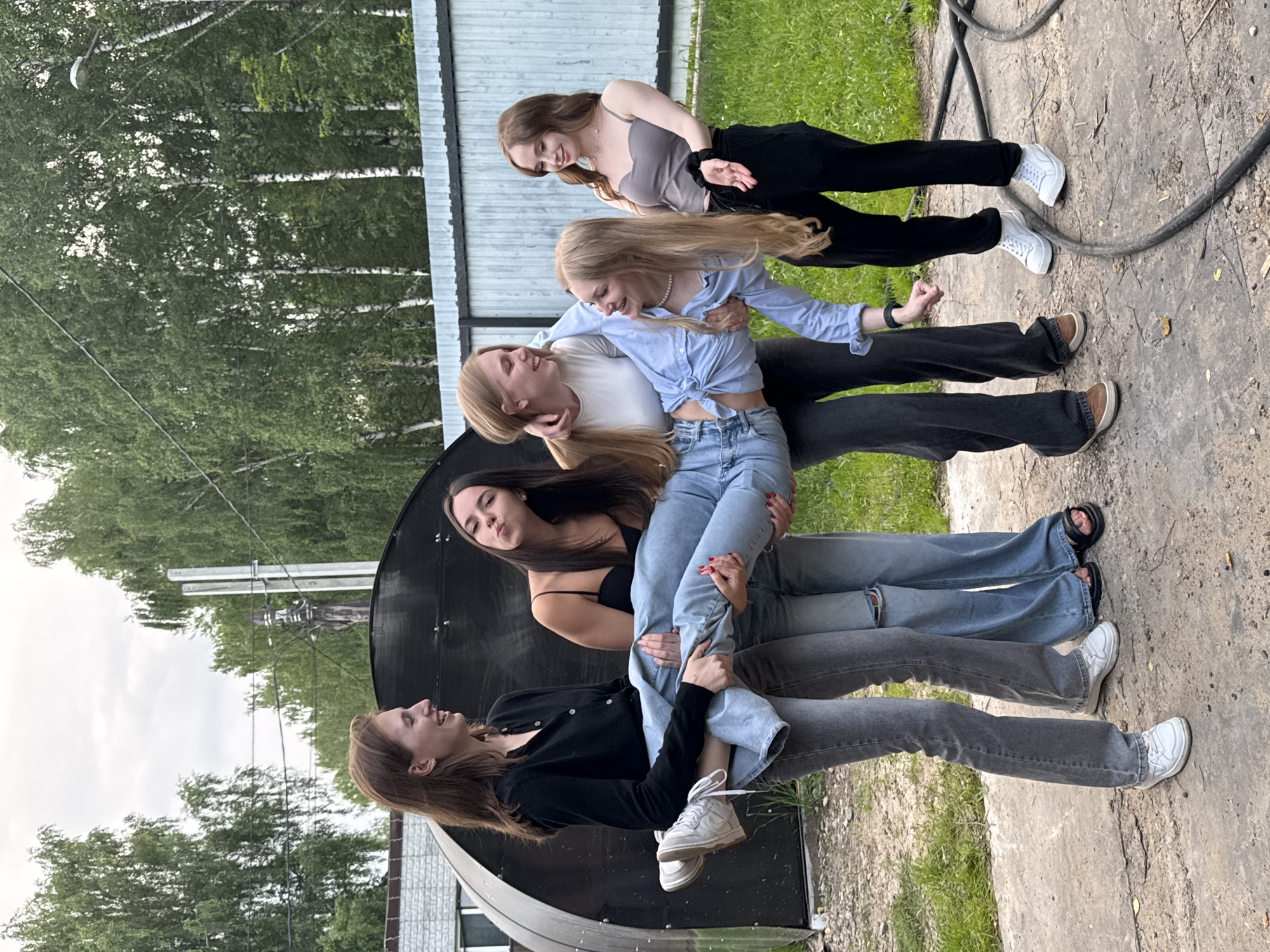 In this photograph, five young women are playfully posing in a line. The image is oriented vertically, making it appear as if the subjects are stacked atop one another, with the topmost woman smiling and looking down and to her right. She is wearing a grey tank top and black pants. Below her, a second woman, with blonde hair, smiles broadly and looks to her left. She is wearing a light blue, tied-up blouse and jeans. The third woman in the stack, with blonde hair also, is looking down and to her left with a slight smile, dressed in a white t-shirt and dark pants. The fourth woman, with long dark hair, is looking down with a pouty expression, wearing a black top and jeans. The bottommost woman, also with long dark hair, is looking up and to her right with a cheerful expression, wearing a black cardigan and grey pants. All the women are wearing casual attire, and their poses suggest a sense of fun and camaraderie.
The setting appears to be outdoors, with a backdrop of green trees and a cloudy sky. A grey, corrugated metal wall or structure is visible behind the women, and a black, curved object, possibly a satellite dish or part of a building, is in the foreground, partially obscuring the lower half of the composition. The ground is a mix of concrete and dirt. A thick black cable runs across the ground in the upper right portion of the image. The lighting suggests it is daytime, possibly overcast. The overall mood is lighthearted and youthful. No visible text is present in the image.