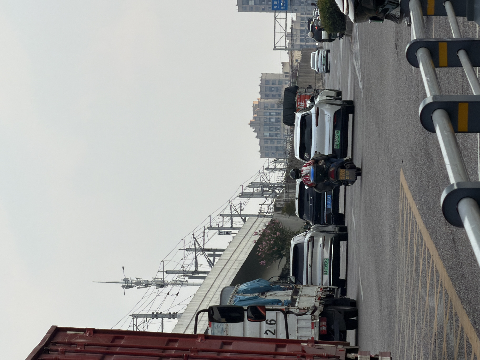 The image depicts a street scene in Jiangjin, China, characterized by moderate traffic flow and urban infrastructure. A large red cargo truck, with the number "26" visible on its side, is positioned prominently on the left, partially obscuring a white truck with blue tarping. In the center of the frame, several cars are in motion, including a silver SUV with license plate "A FX9349" and a black SUV with license plate "G J35A7". A person on a motorcycle, wearing a white and red shirt, is also visible.
The environment includes an elevated road or bridge structure with numerous electrical wires and utility poles overhead, suggesting a well-developed transportation network. In the background, buildings of varying architectural styles are present, with some appearing to be residential or commercial structures. The sky is overcast, indicating a cloudy or hazy day. A railing is present in the foreground, separating the roadway from a pedestrian area or another part of the infrastructure. The overall impression is a typical urban setting during daylight hours.