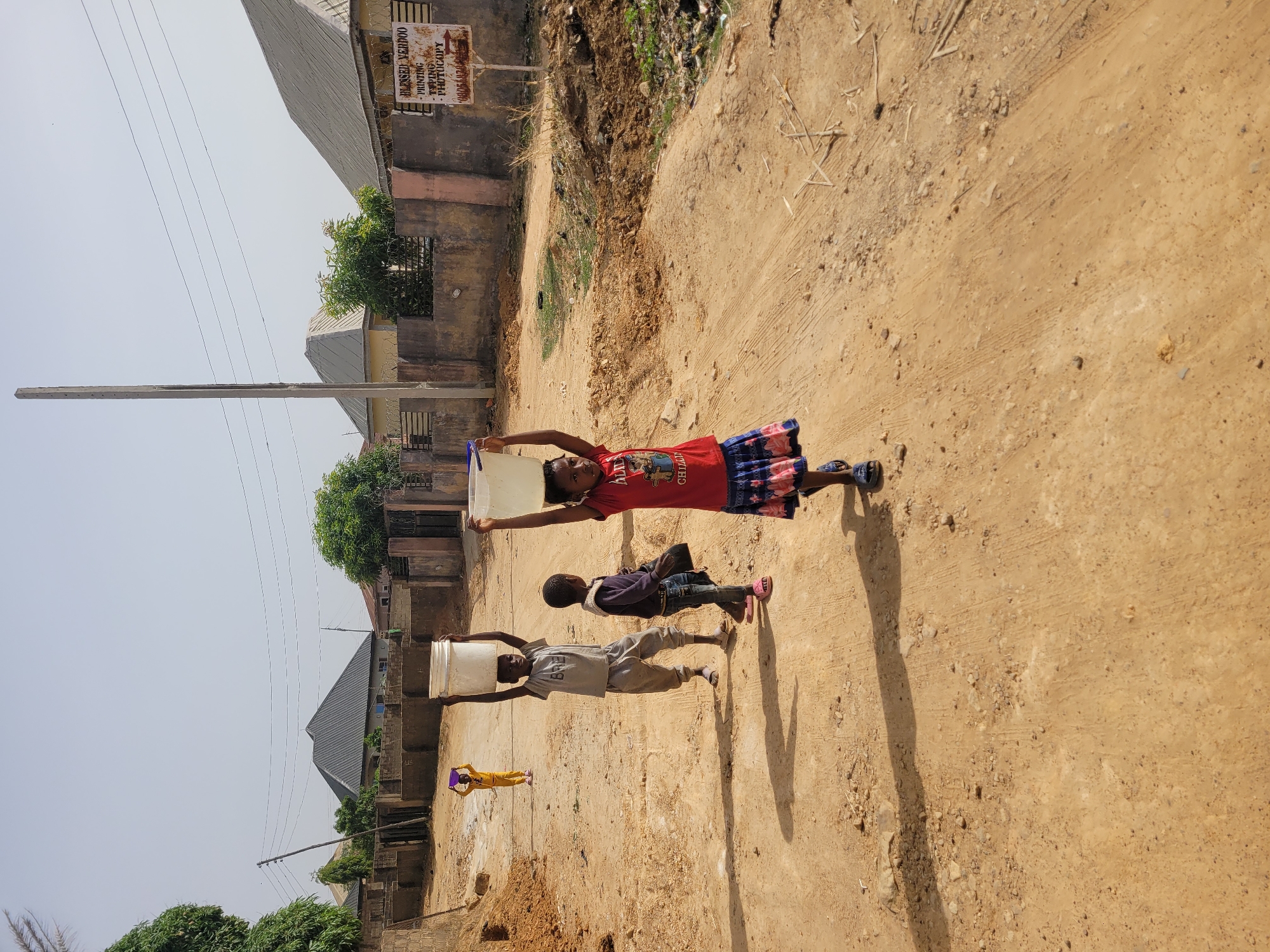 The image depicts a bright, sunny scene on an unpaved dirt road in what appears to be a residential area. Four children are walking along the dusty path, three of whom are balancing containers on their heads, suggesting they are fetching water.
In the foreground, a girl in a red t-shirt and a patterned skirt carries a white plastic bucket on her head, looking towards the viewer. Behind her, a boy in a dark top and jeans walks. Further down the road, another boy in a light-colored t-shirt and pants also carries a white bucket on his head. The smallest child, dressed in yellow, is seen in the distance, balancing a small purple container on her head. Their shadows are short and distinct, indicating strong sunlight, likely around midday or early afternoon.
The road is bordered by simple buildings with concrete walls and corrugated iron roofs, interspersed with some greenery. A tall utility pole with overhead electrical wires is visible on the left. On the right, a brown sign is attached to a building, displaying the text: "BLESSED YERDOO", followed by services "- PRINTING", "- TYPING", "- PHOTOCOPY", and a partially visible phone number starting with "08054084". The scene captures daily life in Barakin Akawo, Nigeria, where manual water collection is common.