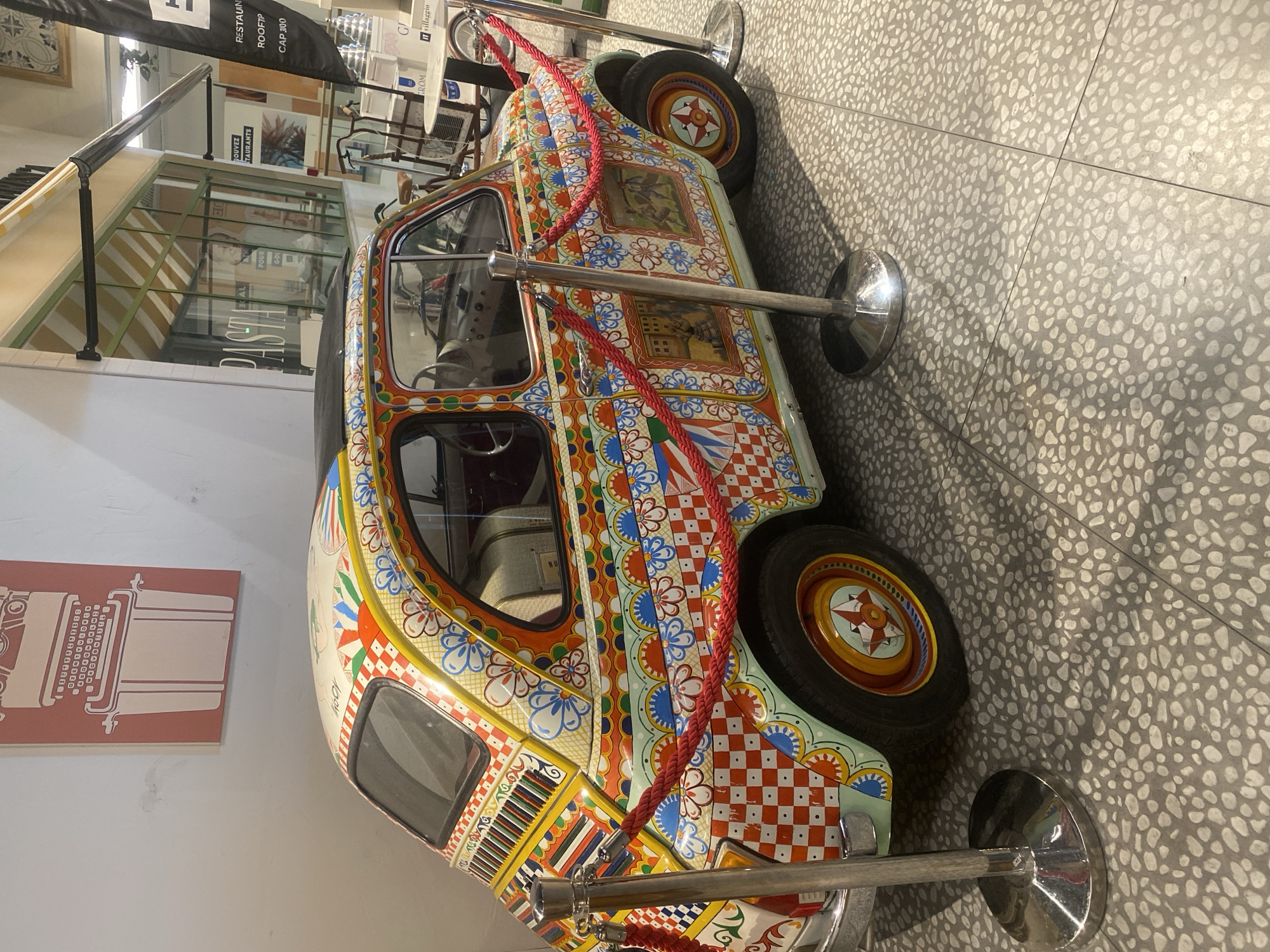 The image shows a brightly decorated Fiat 500 parked indoors, possibly in a shopping mall or exhibition space in Saint-Laurent-du-Var, France. The car is adorned with vibrant, intricate patterns, including floral motifs, geometric shapes, and small painted scenes. Red ropes are set up around the vehicle, indicating it is on display and not accessible to the public. The flooring is a speckled terrazzo pattern. In the background, a large print of a vintage typewriter hangs on a white wall. Further in the background, a storefront with a striped awning displays the word "PASTA" and other text in French. Other objects visible in the background include bicycles, tables, and flags.