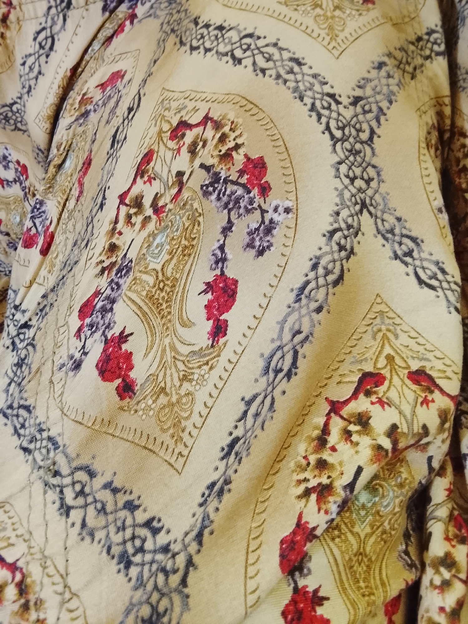 The image shows a section of fabric with a complex, patterned design. The dominant color is a light, creamy beige. The pattern consists of repeating motifs arranged in a grid-like fashion. These motifs include floral elements featuring red and brown flowers, and ornate, baroque-style ornamental shapes with tan and brown detailing. Separating the motifs are dark gray geometric borders with curved elements. The fabric appears slightly wrinkled. The image does not depict any specific location or actions.