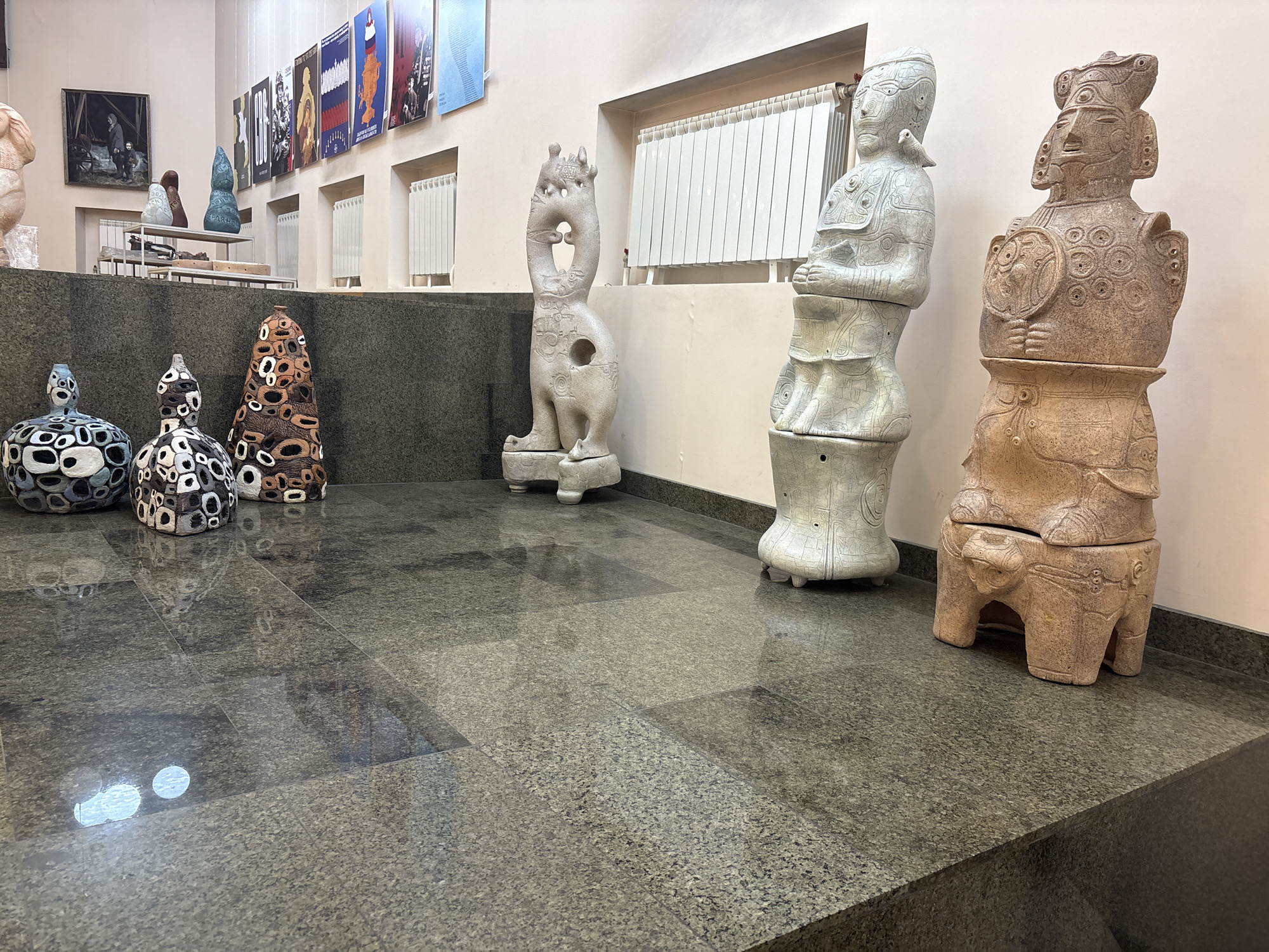 The image captures an indoor space, potentially a museum or gallery, showcasing a collection of art pieces. On the left, a group of spherical sculptures with contrasting black and white patterns, along with another with brown and white patterns, are positioned. Moving towards the center, there is a tall, elongated sculpture of a mythical creature, followed by two larger, anthropomorphic sculptures. These stand near a window with a radiator. The room has a polished, reflective floor, with posters or artworks hanging on the wall in the background. The lighting appears to be artificial.