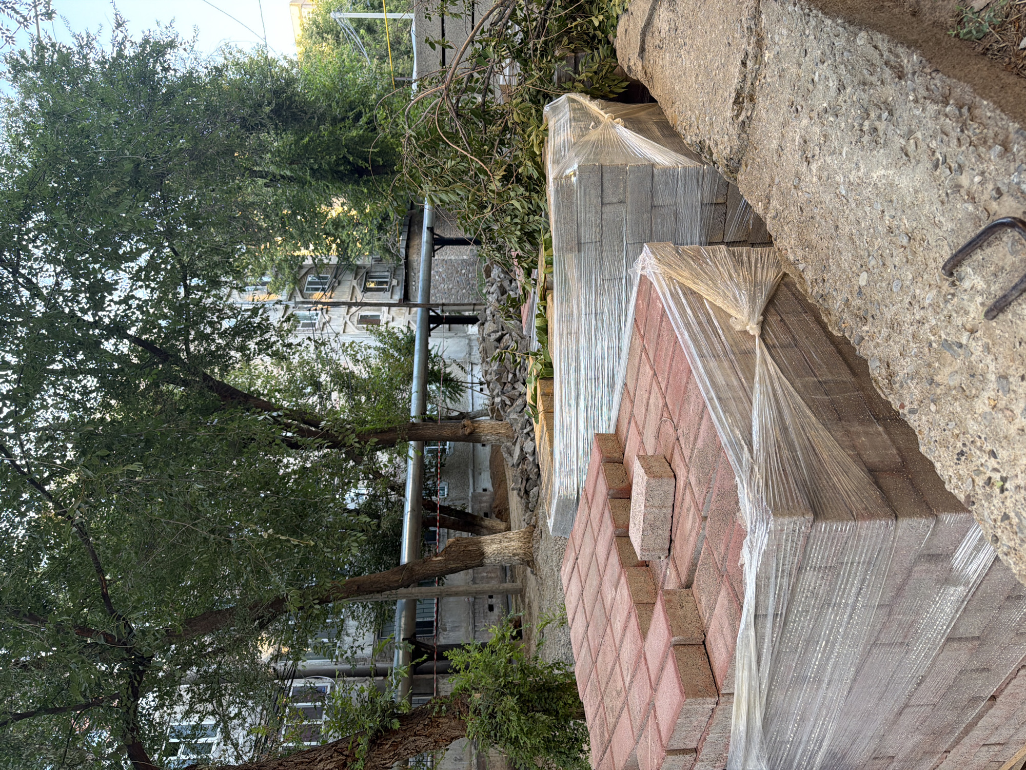 In Shymkent, Kazakhstan, an outdoor scene depicts construction materials piled in front of a residential building. In the foreground, stacks of red and grey paving stones are neatly arranged, some covered in clear plastic wrap. A concrete curb runs along the right side of the image, with exposed rebar. Behind the paving stones, a pile of broken concrete and rocks is visible. Trees with green foliage partially obscure a multi-story building with numerous windows. A large pipe runs horizontally across the middle ground, supported by posts. The ground is covered in dirt and gravel. The sky is clear and blue.