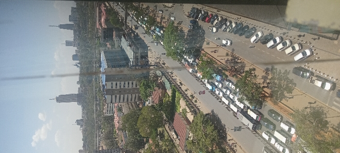 An elevated, wide-angle image captured in daylight depicts a bustling urban scene in Nairobi, Kenya, looking northeast towards the Central Business District and Upper Hill. The vantage point is from a high-rise building, suggested by the vertical element in the center-right of the frame, likely a window mullion.
In the foreground, two primary parallel roadways are visible. The left-hand roadway features a continuous queue of vehicles, primarily white and light-colored passenger cars, SUVs, and multiple minibuses (matatus), indicating heavy traffic congestion as vehicles proceed towards the distant city center. The right-hand roadway shows a mix of moving vehicles and numerous cars parked diagonally and parallel along its left curb, predominantly white. Sidewalks border both roads, populated by indistinct pedestrians.
The mid-ground contains a mix of multi-story buildings of varying architectural styles and significant green spaces with mature trees. A prominent grey multi-story building with numerous windows is situated on the left side of the main left roadway. An elevated highway or bridge structure is visible spanning across the mid-ground, beneath the distant skyline.
The background features the distinct skyline of Nairobi. Key identifiable landmarks include the Britam Tower, a tall, slender skyscraper with a unique apex, situated on the far left of the distant skyline. To its right, the Kenya International Convention Centre (KICC) tower, characterized by its distinctive circular top, is visible. Numerous other high-rise office and residential buildings constitute the dense urban landscape of the Central Business District and Upper Hill.
The overall environment suggests a clear day with scattered clouds and strong sunlight, indicating midday or early afternoon. The scene is characterized by active vehicular traffic, parked cars, and pedestrian movement in a major urban corridor. Given the visible landmarks and general orientation, the image was captured from an elevated position within the **Kilimani** or **Upper Hill** area of Nairobi. The main road with heavy traffic in the foreground is likely a section of **Valley Road** or **Uhuru Highway** approaching the city center.
