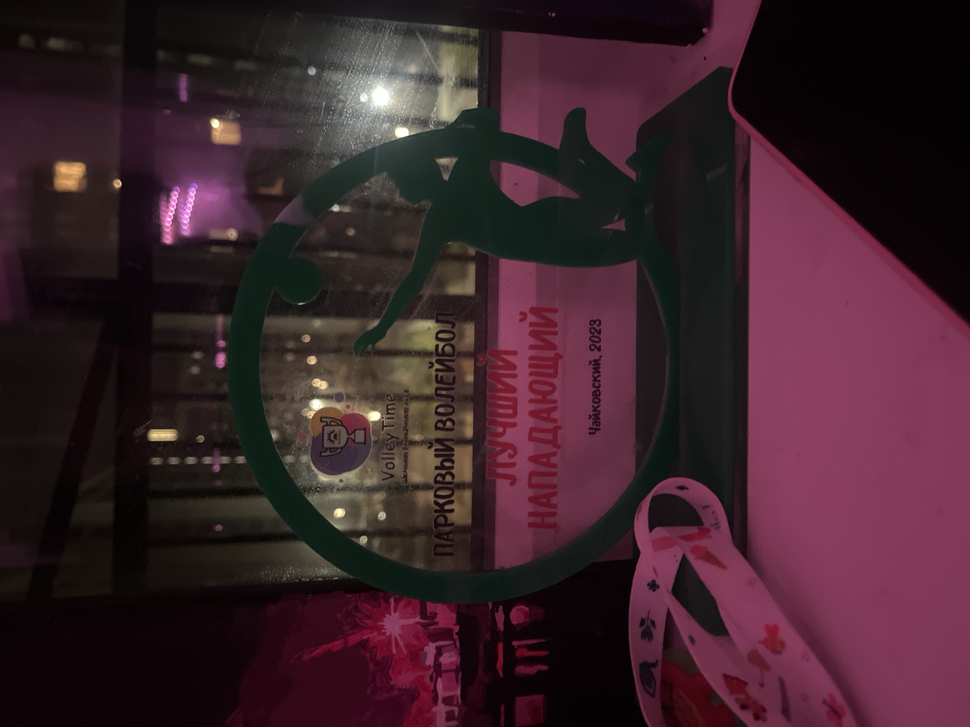 The image displays a translucent green award or trophy, shaped as a circular frame enclosing the silhouette of a volleyball player in an attacking pose. The award is placed against a window, revealing a dark night cityscape in the background, characterized by numerous blurred lights creating a bokeh effect. A prominent pinkish-purple light source or reflection illuminates parts of the scene, including the award and the adjacent wall.
Text written in Cyrillic is visible on the award. It reads: "ПАРКОВЫЙ ВОЛЕЙБОЛ" (Park Volleyball), "ЛУЧШИЙ НАПАДАЮЩИЙ" (Best Attacker/Spiker), and "ЧАЙКОВСКИЙ, 2023" (Tchaikovsky, 2023). A smaller logo at the top left of the award includes the text "Volley Time" and partially visible words that likely say "Школа пляжного волейбола" (School of beach volleyball).
To the right of the award, a white fabric ribbon is visible, adorned with small, colorful patterns such as hearts, flowers, and ice cream cones. A dark electronic device, possibly a smartphone or tablet, is partially visible along the right edge of the frame. The scene captures a personal memento, indicating an achievement in volleyball, likely set indoors with a view of a city at night. The overall atmosphere is dim, highlighted by the glowing city lights and the distinct pink hue. This scene is contextualized as being in Saint Petersburg, Russia.