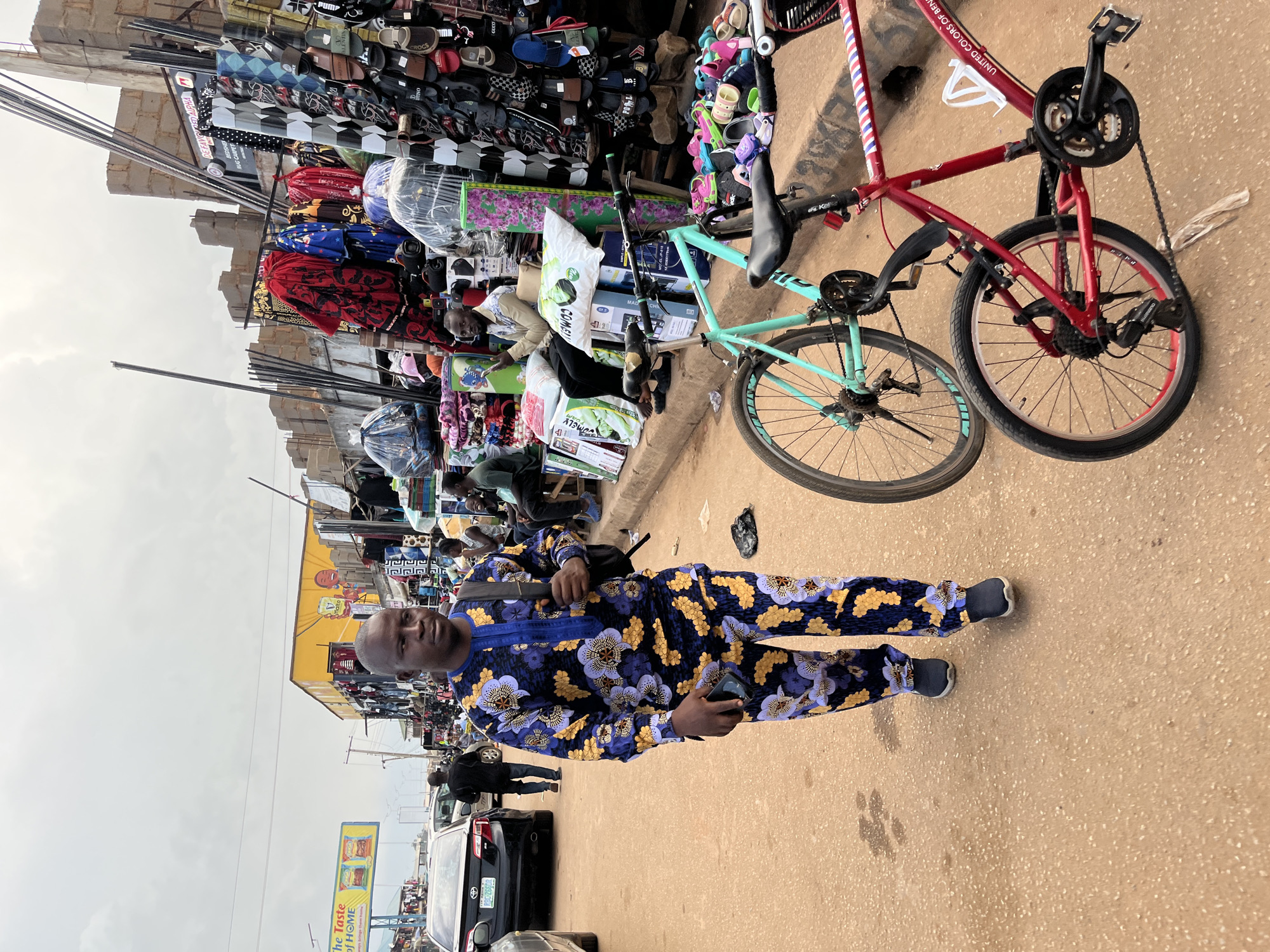 This image captures a street scene in Bukuru, Nigeria, with a focus on a man walking towards the viewer. He is wearing a vibrantly colored traditional African outfit with a blue base and a bold floral pattern in yellow and purple. He carries a black backpack and holds a mobile phone in his right hand. The street itself is paved with a reddish-brown earth-colored material, showing signs of wear and tear with some scattered debris.
To the left of the man, a black sedan car is parked, and further down the street, other cars and pedestrians are visible, suggesting a bustling urban environment. On the right side of the image, a market stall is set up, displaying a variety of goods, including racks of footwear, textiles, and other assorted items. Two bicycles are also present in the foreground: one is a light teal color, and the other is a red frame bicycle with a white water bottle holder. The background shows a mix of market structures, unfinished buildings, and overhead wires, under a cloudy sky. The overall atmosphere appears to be that of a busy marketplace in Nigeria.