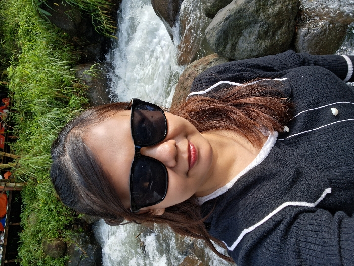 A woman wearing dark sunglasses and a black sweater with white stripes and pearl accents poses in front of a waterfall. Lush green vegetation grows nearby, and the river water cascades over rocks. The woman's hair is brown and layered. Her lips are painted a reddish hue, and she appears to be smiling slightly. The scene appears to be taken outdoors on a sunny day.