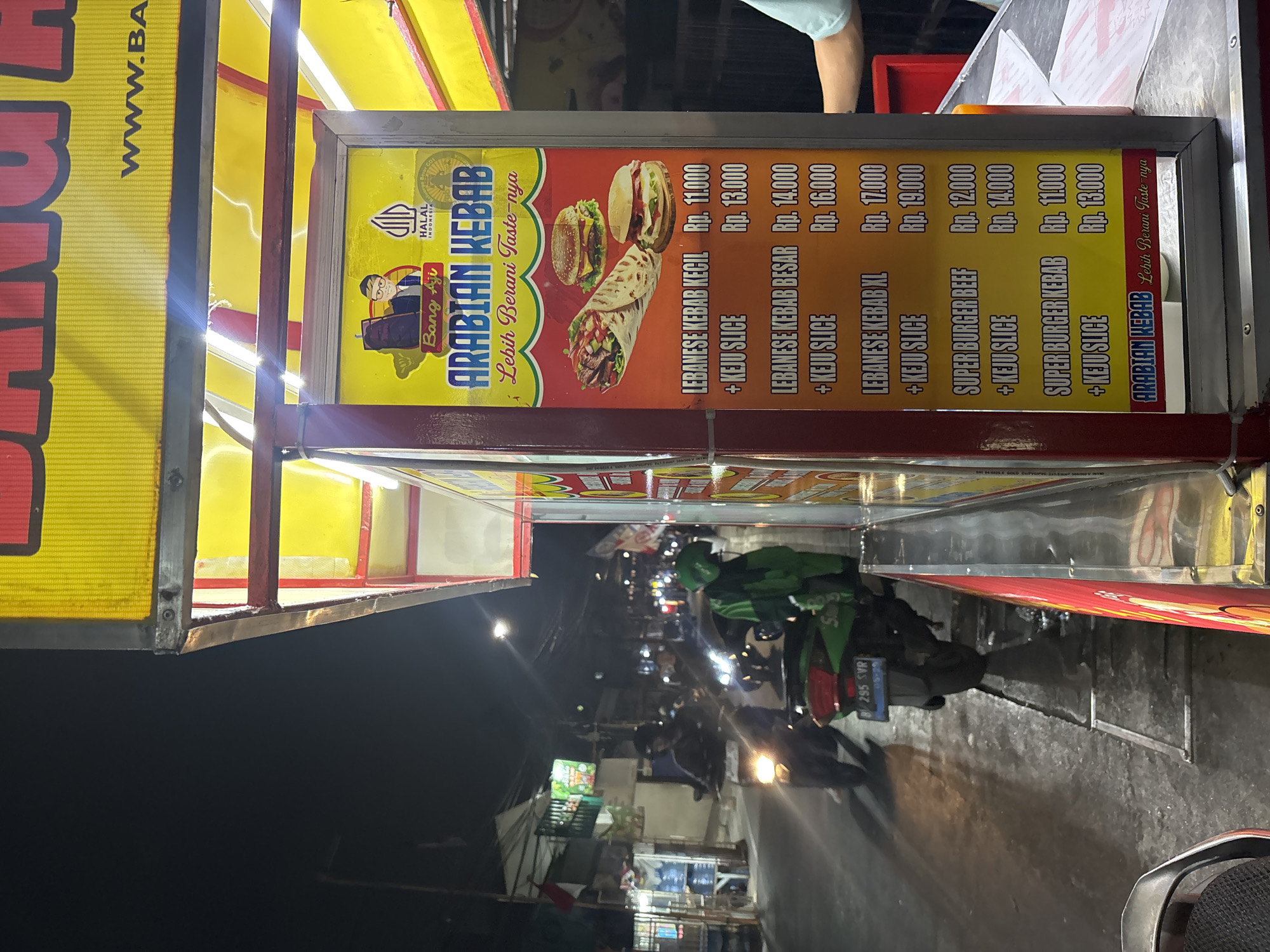 A night-time street scene in North Jakarta, Indonesia, shows a brightly lit food stall selling Arabian Kebab. The stall features a large, colorful menu board displaying various kebab and burger options with prices listed in Indonesian Rupiah. Two types of kebabs, "Lebanese Kebab Kecil" and "Lebanese Kebab Besar," are pictured along with two burgers. The stall also offers "Super Burger Beef" and "Super Burger Kebab." The menu board is illuminated by a yellow canopy above. In the background, a street scene unfolds with several motorcycles, one of which has a rider wearing a green jacket and helmet, possibly a delivery driver. Streetlights illuminate the road, and buildings line the street. The overall impression is of a bustling street food environment.