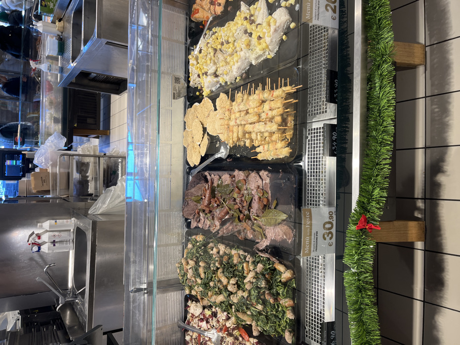 This media file captures a deli or prepared food counter in Rozzano, Italy, likely during the holiday season. The central focus is a refrigerated glass display case showcasing various pre-prepared dishes in black trays.
The visible dishes, from top to bottom, include:
* A white, creamy salad-like dish topped with yellow and dark ingredients, possibly potatoes or corn and herbs.
* Numerous skewers, likely shrimp or chicken, alongside breaded or fried items.
* Slices of roasted meat, such as roast beef, garnished with bay leaves and other seasonings.
* A green salad featuring mixed greens and white protein chunks, possibly chicken or cheese.
* A salad containing red radicchio and other ingredients.
Alongside the food, several white pricing labels are visible. One prominent label states "Menú delle Feste" (Menu of the Festivities/Holidays) with "PRODUZIONE PER" (Production for) and a price of "€30,90". Other clearly visible prices include "€34.90", "€33.90", and a partial "€20". A festive green garland adorned with a small red bow is attached to the side of the display case, reinforcing the holiday theme.
The background reveals a commercial kitchen area with stainless steel countertops, a sink with a faucet, cleaning product bottles, and industrial kitchen equipment. Reflections of people and the indoor environment are faintly visible on the glass and metal surfaces, suggesting customer or staff presence. The scene depicts a bustling food service environment offering ready-to-eat meals, possibly for festive occasions.