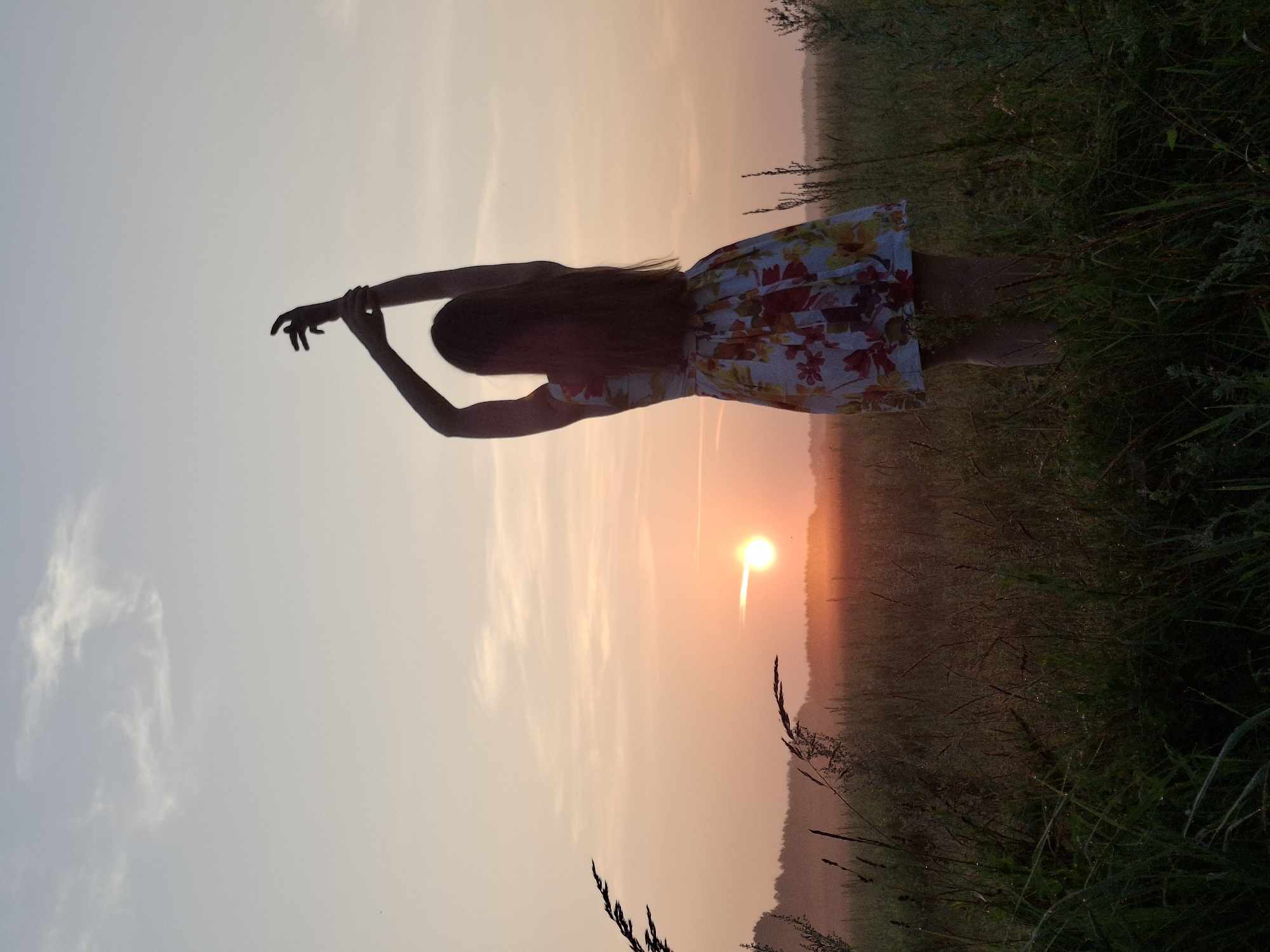 A full-body, silhouette shot captures a young woman standing in a field at sunset. She wears a floral sundress and has her arms raised above her head, with her hands clasped. The sun is setting on the horizon, casting a warm orange glow across the sky. The field is filled with tall grass and wildflowers, and a line of trees can be seen in the distance. The overall mood of the image is peaceful and serene. The location is identified as Plishkino, Russia, though no specific landmarks are visible to confirm this.