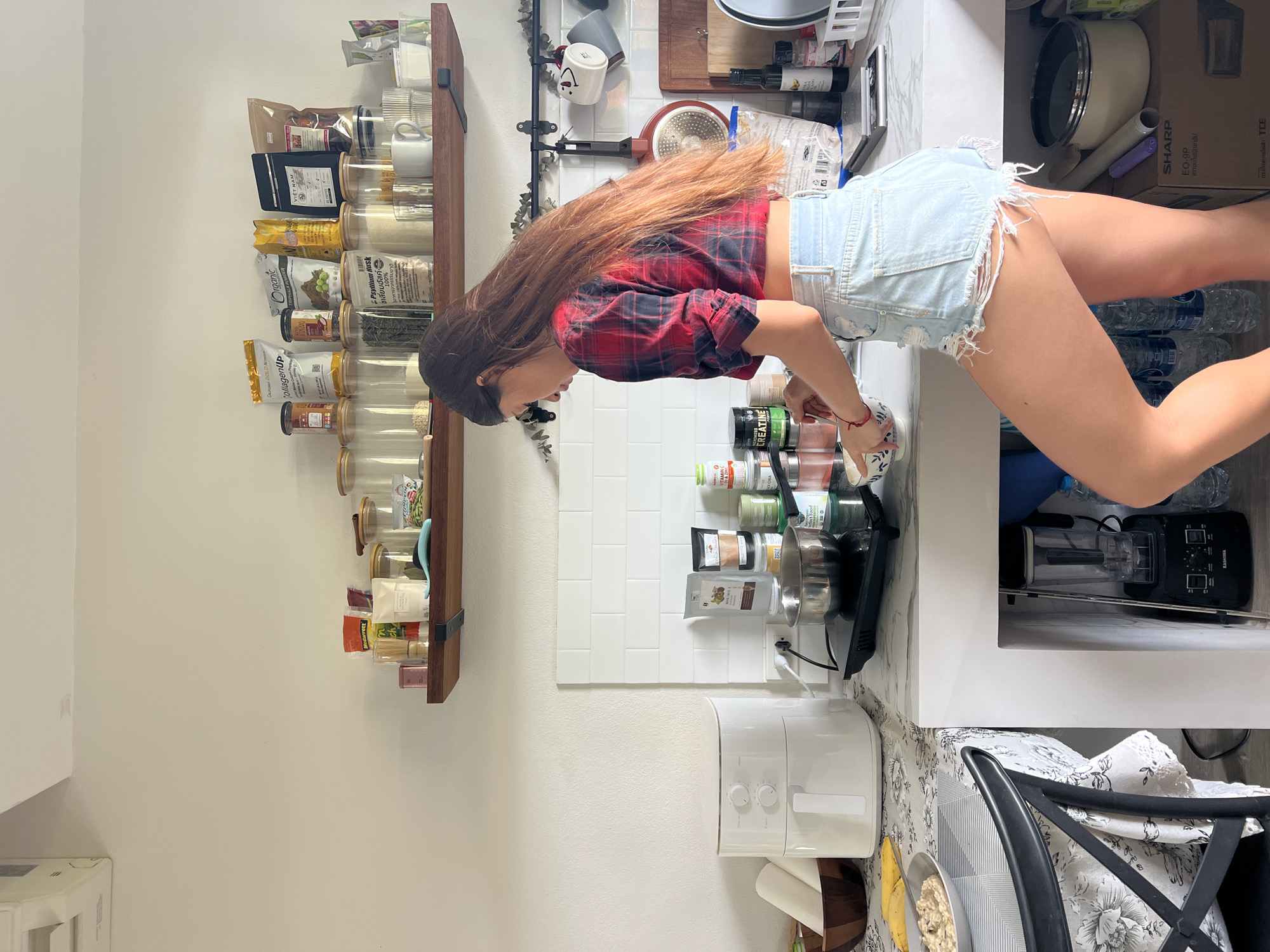 A waist-up shot shows a woman standing at a kitchen counter, possibly in a home kitchen in บ้านบางรักษ์, Thailand. She is wearing a red and black plaid shirt and denim shorts. Her long brown hair flows down her back. The woman appears to be interacting with items on the counter, possibly preparing food. The counter is lined with various objects, including a blender, spices, jars of food items, and a large white appliance (possibly an air fryer). A shelf on the wall behind her displays more food items in jars. In the background, there are water bottles and a box labeled "SHARP" indicating an appliance. A chair and a bowl with bananas are visible in the lower right corner.
