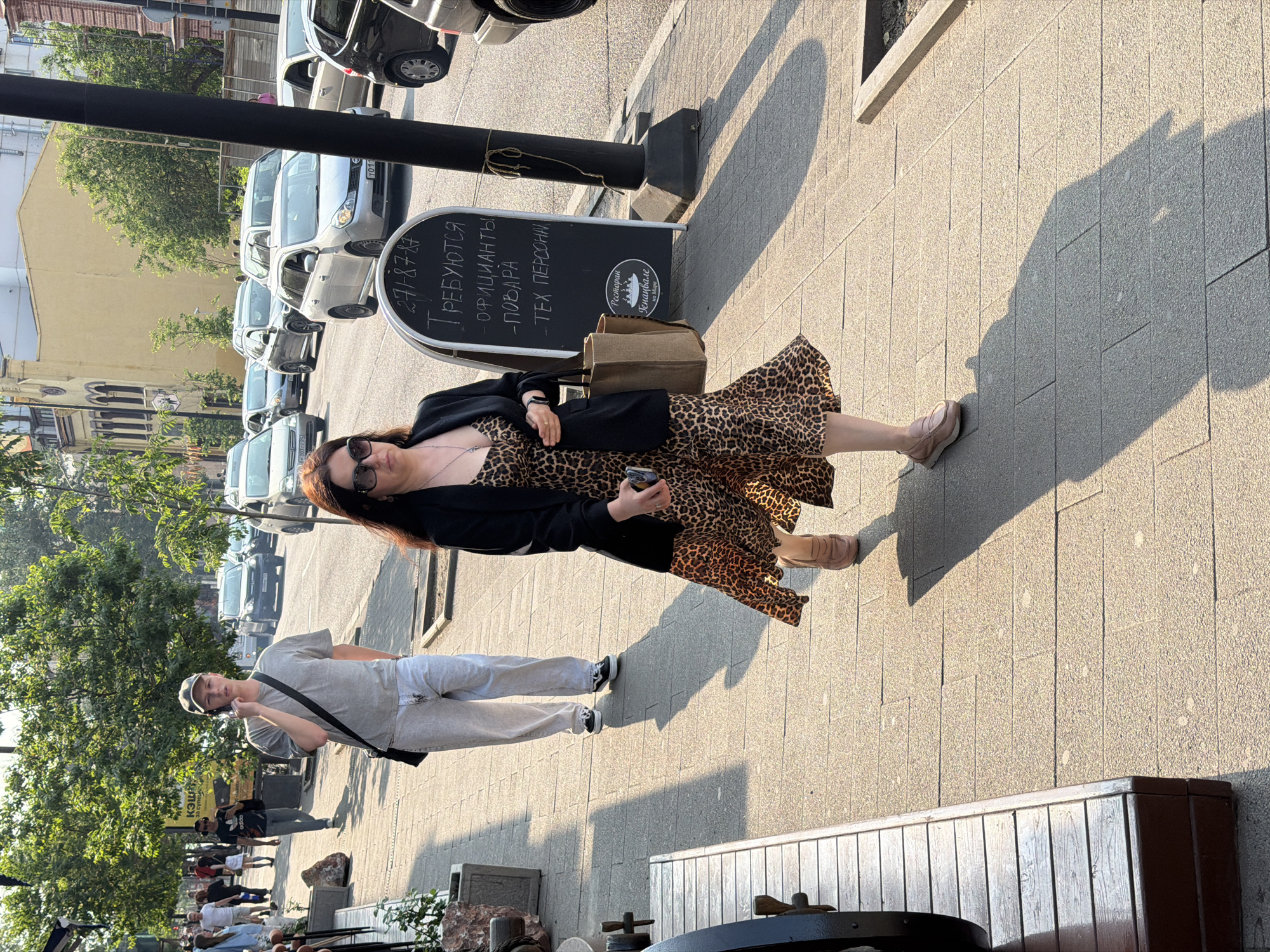 A woman wearing a leopard print dress and a black blazer walks along a sidewalk in Krasnoyarsk, Russia. She is holding a smartphone in her right hand and a large tote bag in her left. A man in a grey t-shirt and a baseball cap is walking in the background, also on the sidewalk, and talking on his phone. Cars are visible on the street behind them. A blackboard sign with text in Cyrillic is placed on the sidewalk to the right of the woman. The scene appears to be a sunny day.