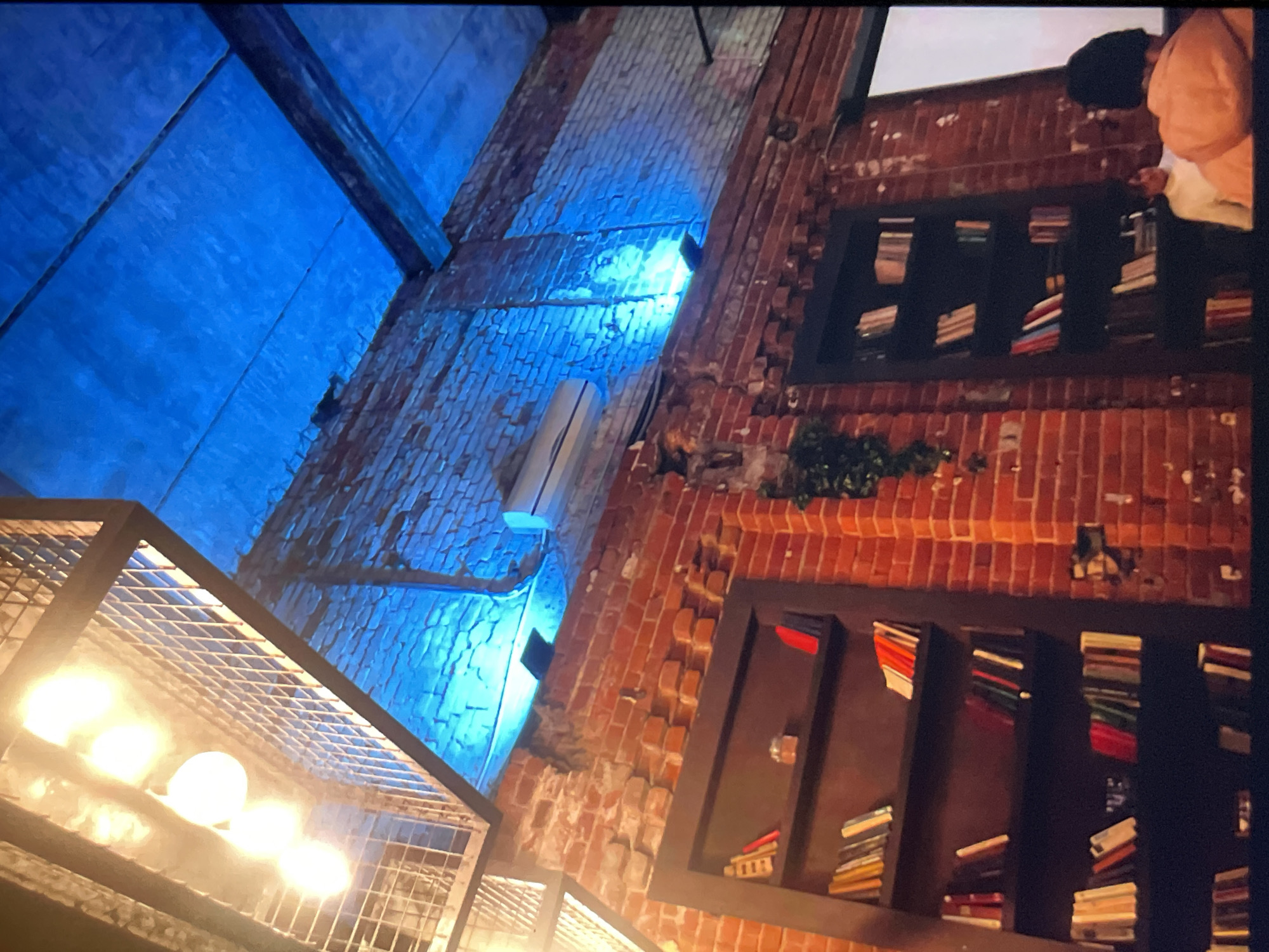 Here is a detailed description of the image:
The image shows an interior space with exposed brick walls. Two black bookshelves are recessed into the brick, filled with stacks of books. There is a person partially visible on the right side of the upper bookshelf, wearing a light-colored top. A hanging air conditioning unit is mounted on the brick wall between the bookshelves. Below the bookshelves, there's a caged lighting fixture with illuminated bulbs. The upper section of the wall is illuminated with blue light, suggesting a window or skylight.