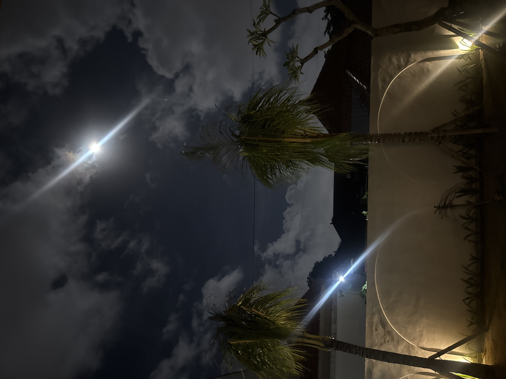 This nighttime image, possibly taken in North Kuta, Indonesia, features a view of the sky with clouds and a bright celestial object, likely the moon. Two palm trees are in the frame. One is in the upper right corner, and the other is in the lower left corner. A building and landscape elements are visible in the background, illuminated by a light source. There is a wall with a bright light on it.