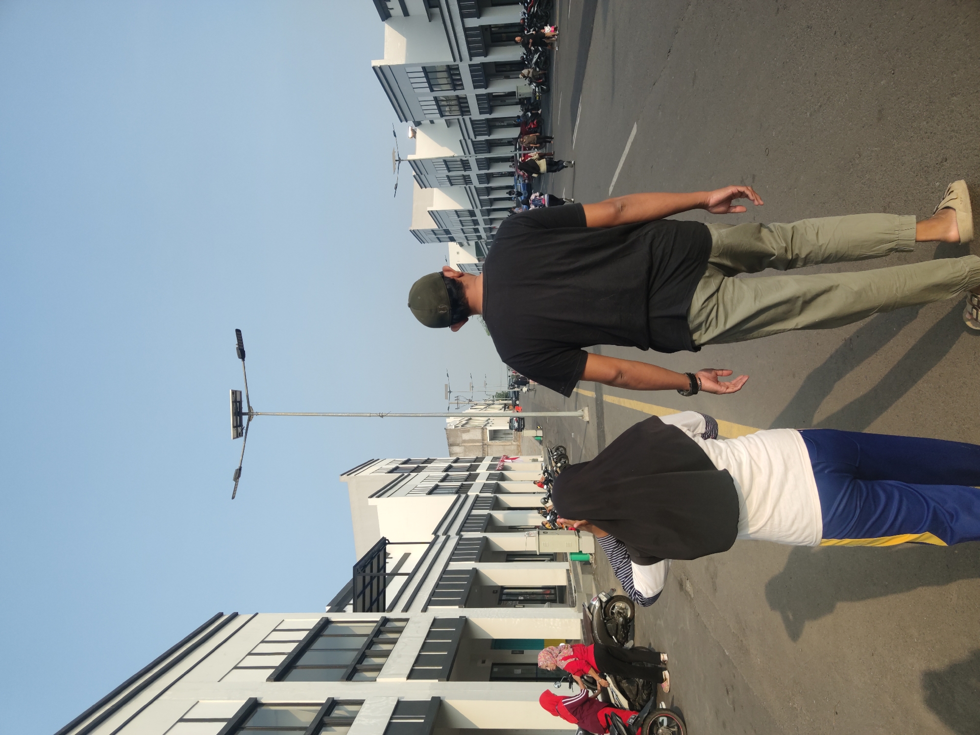 The image depicts a street scene in Petiken, Indonesia, with a clear sky and sunlight casting shadows. Two individuals, a man and a girl, are walking away from the camera on a paved road. The man, wearing a black t-shirt and olive-colored pants, has a dark green cap on his head. The girl, seen from behind, is wearing a black hijab, a white long-sleeved shirt, and blue and yellow striped pants.
On the left side of the road, a row of modern, white buildings with black trim and large windows lines the street. Several motorcycles are parked along the sidewalk in front of these buildings. In the foreground on the left, two women wearing hijabs are standing near a parked motorcycle. One is wearing a red hijab and a red long-sleeved shirt with dark pants, while the other has a pink hijab and a red shirt with dark pants.
Further down the road, more buildings of a similar architectural style are visible, stretching into the distance. Other people and motorcycles can be seen scattered along the street and sidewalks. A tall street lamp with solar panels stands near the center of the frame. The road has a yellow dividing line.