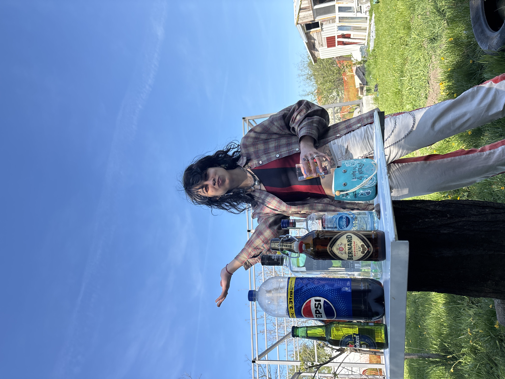 A person with long dark hair stands behind a white table filled with various bottles and containers under a bright blue sky. The individual wears a plaid shirt over a striped top and light-colored pants with a red stripe down the side. They hold a glass in one hand and gesture towards the bottles with the other. The bottles include brands such as Pepsi, Bernard beer, and Poliakoff vodka, among others.
Behind the table and person, there is a grassy area and a dilapidated structure with a collection of windows. A tire leans against the structure on the right side of the frame.
Based on the label "39€/3.31лв" and the Cyrillic lettering on some of the bottles (e.g., "Банкя"), the location is likely in Bulgaria. The text "Bernard" is also present on multiple bottles.