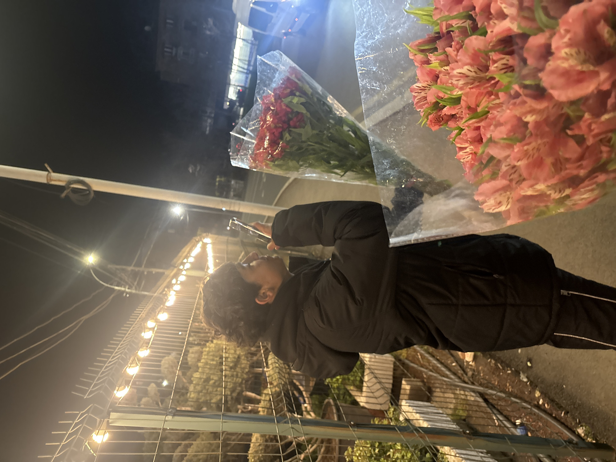 A person in a dark winter jacket and black pants stands at a flower stand at night, looking at their phone. They stand near a metal fence decorated with string lights. Two flower arrangements are displayed: one with red flowers and the other with pink. The scene is outdoors, on a street in Yerevan, Armenia.