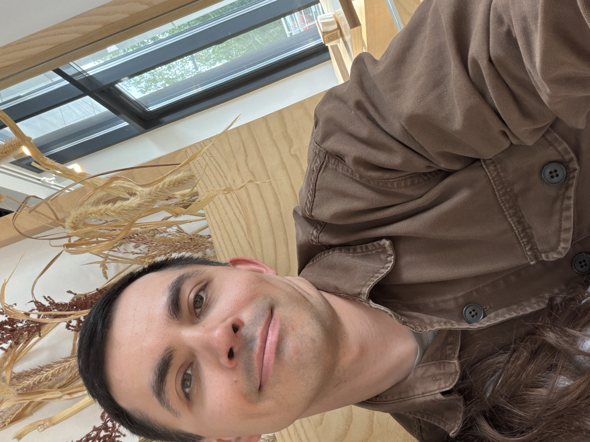 A man is taking a selfie indoors. He is wearing a brown button-up shirt. Behind him are decorative dried plants and a window. The location is unknown, but is possibly a cafe.