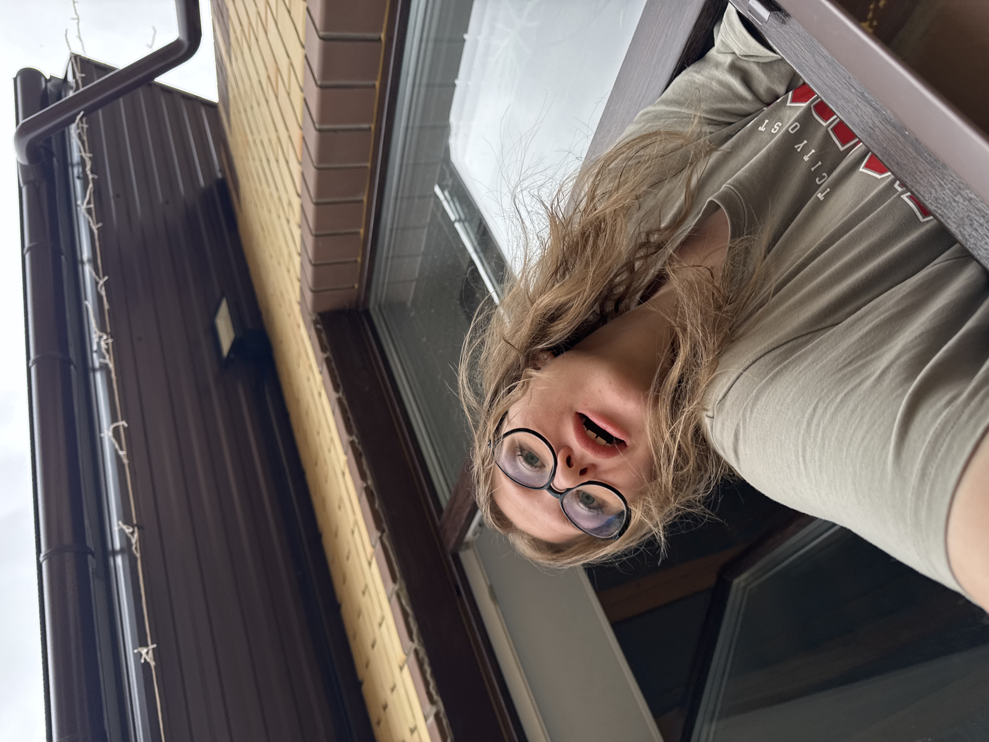 A low-angle, outdoor shot depicts a person looking down into the camera from a building opening, likely a window. The subject, a young individual with long, wavy light brown hair, wears black-rimmed glasses and a light grey t-shirt. Their mouth is slightly open, and their gaze is directed downwards, suggesting curiosity or surprise. Partially visible red and white text on the t-shirt reads "CITY OF ST".
The background on the left side of the image features the exterior of a building, comprising sections of light brown brickwork and dark brown corrugated metal siding. A prominent dark brown gutter and downspout run vertically along the building's facade, adorned with a string of white fairy lights. The sky above is bright and overcast. The right side of the frame shows a dark window frame and the darker interior space from which the person is observing. The image captures an overcast daytime scene in Gornyi (Dvurechenskoe S/P), Russia.