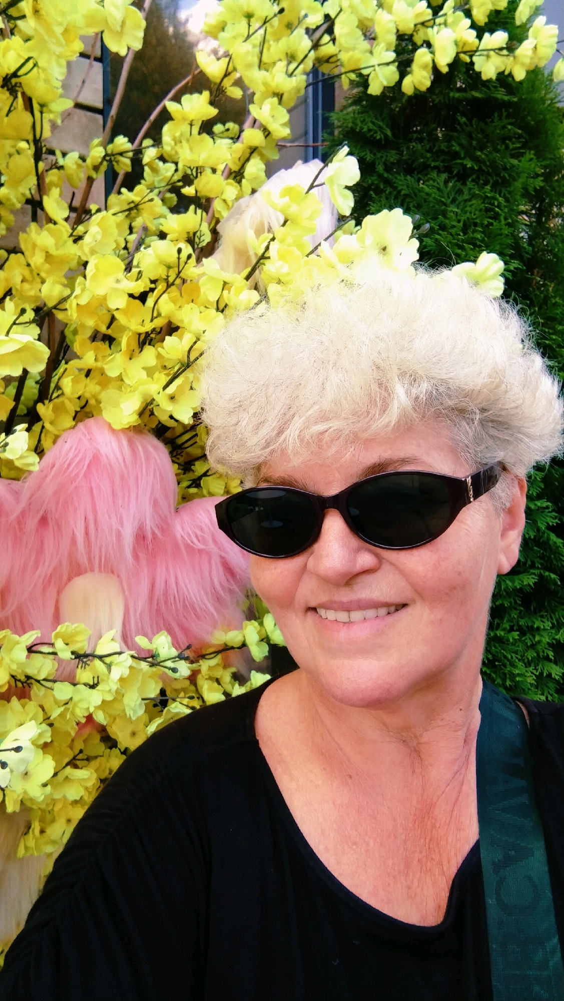 A woman with blonde, curly hair and dark sunglasses smiles at the camera, standing in front of a display of yellow artificial flowers and a pink fluffy object. She is wearing a black t-shirt. The background includes a dark green evergreen tree and hints of a building facade behind the flowers. The scene appears to be an outdoor display, possibly in a public space or shop in Horishni Plavni, Ukraine.