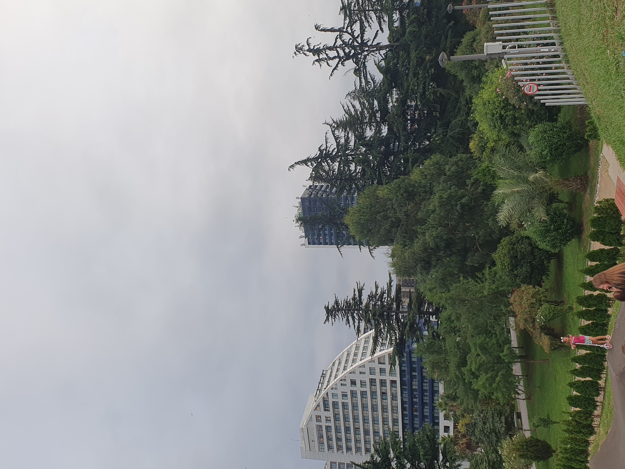 In the city of Batumi, Georgia, a cloudy day is captured in a wide-angle shot that presents a modern architectural complex and a vibrant green park. The left side of the image is dominated by a striking, angular white building with a curved, stepped facade, possibly a hotel or residential complex. Its numerous windows are visible, reflecting the overcast sky. To the right, a darker, taller building, also modern in design, rises above a dense line of tall evergreen trees, likely pine or cedar, with their characteristic feathery foliage.
In the foreground, a lush park area unfolds. A young girl, dressed in a pink top and shorts, is captured in motion on a pink scooter, her body angled slightly as she rides. Her hair is adorned with a flower accessory. Behind her, a path winds through the park, lined with neatly trimmed green bushes. Further into the park, various trees and shrubs create a rich tapestry of green hues, with some flowering bushes adding touches of red and pink. A white metal fence runs along the right edge of the park, demarcating its boundary. A lamppost and a white weather station are visible near the fence, along with a circular red sign with white lettering, the contents of which are illegible due to the distance and resolution. The ground in the immediate foreground is a darker, textured surface, possibly a pathway or a play area, with a hint of red. Two people are partially visible in the lower right corner, their faces turned away from the camera, suggesting they are engaged in conversation or observing the park. The sky above is a uniform pale gray, indicative of cloudy weather.
