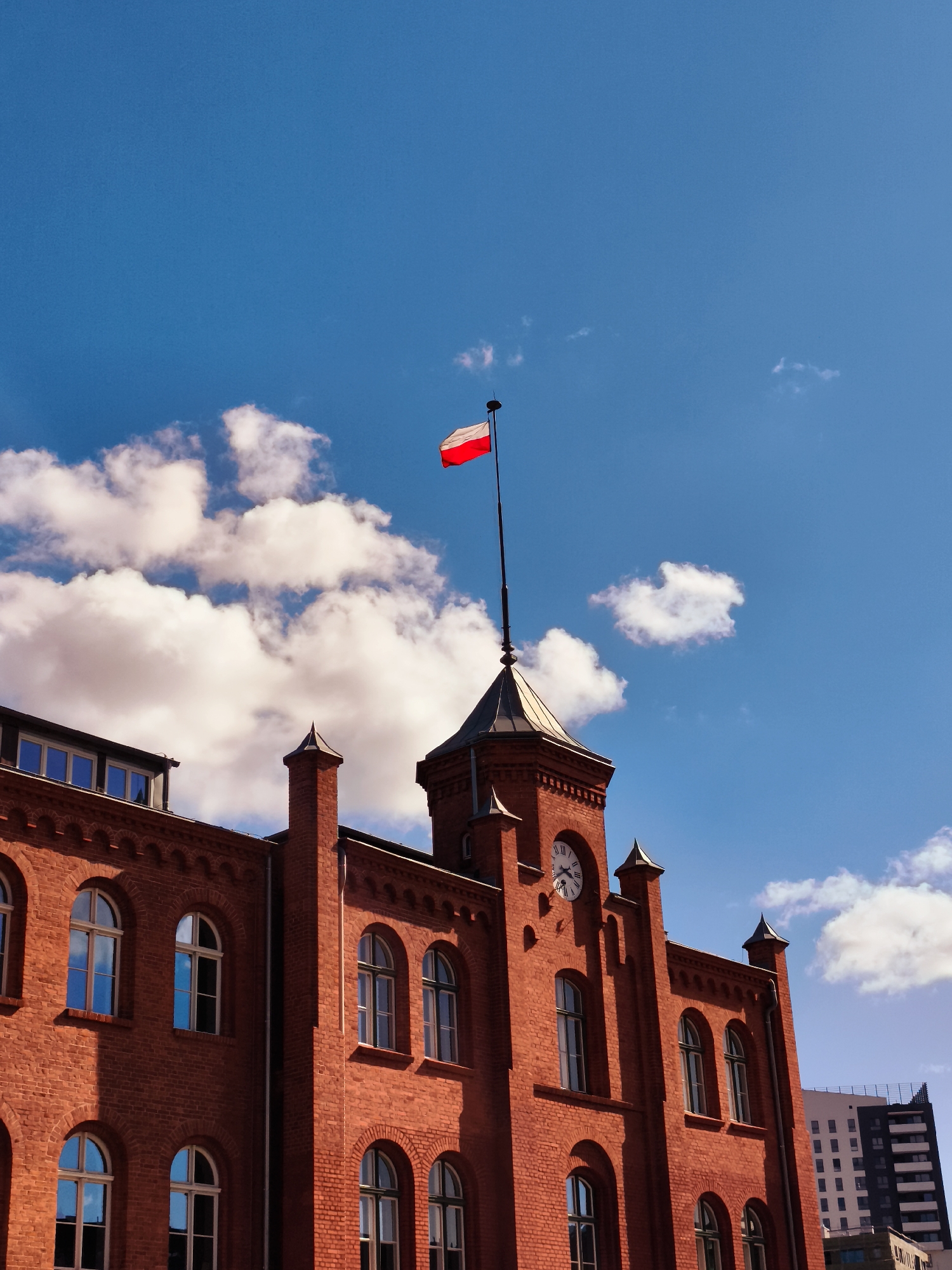 The image shows the exterior of a historic red brick building in Gdańsk, Poland. The building features arched windows and a prominent clock tower. A Polish flag flies from a flagpole atop the tower, silhouetted against a bright blue sky with scattered white clouds. The scene captures a section of the building's facade and the sky above, suggesting a clear day.