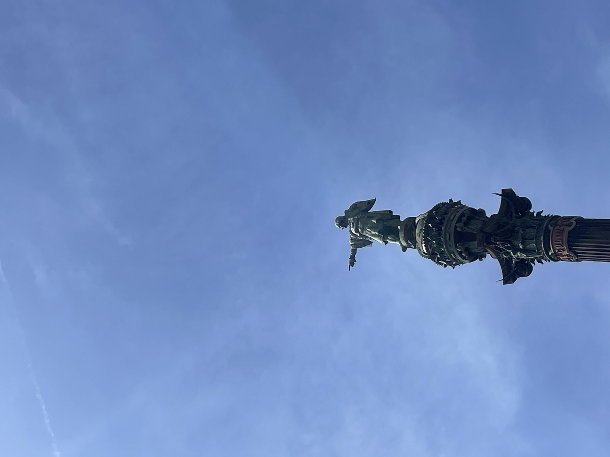 The image shows a low-angle view of the Columbus Monument in Barcelona, Spain, against a clear blue sky. The statue of Christopher Columbus stands atop a highly ornamented column, pointing away from the city. The column's base and shaft have intricate carvings and architectural details. The sky is mostly empty except for contrails.