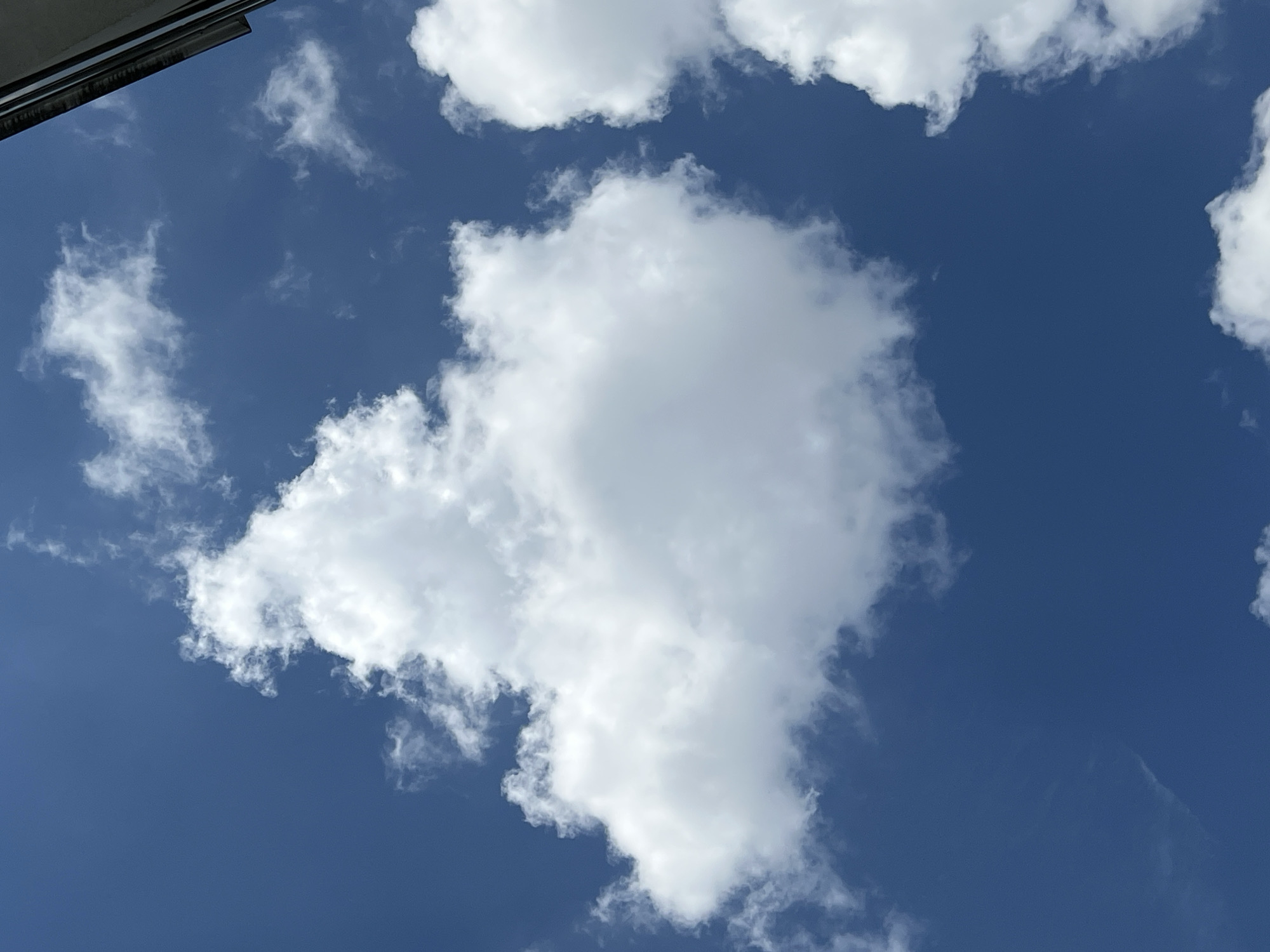 The image shows a section of the sky with white clouds against a blue background. A small portion of a building structure is visible in the upper left corner. There are no people or discernible actions taking place. The environment is open sky. The image does not contain sufficient information to determine the exact location within the city of Krasnodar, Russia.