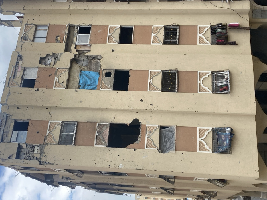 A multi-story residential building in Khan Yunis, Palestinian Territory, exhibits extensive damage from apparent conflict. The facade, primarily beige with brown vertical accent panels and ornate, light-colored arch-shaped frames above many window spaces, is heavily compromised. The upper two floors show severe structural collapse, with large sections of the exterior missing, exposing bricks, concrete, and twisted rebar. Numerous circular impact marks and perforations are visible across the entire facade, consistent with bullet or shrapnel damage.
Multiple windows are completely absent or shattered, leaving dark, empty openings. Large, irregular-shaped impact craters penetrate several floors, exposing interior spaces. On the fourth floor from the bottom, a prominent blue tarpaulin covers a window opening in the central column. Another grey sheet covers an opening on the second floor from the bottom, also in the central column. Several windows retain metal security grates, some of which appear bent or damaged. On the bottom right floor, various pieces of clothing are seen hanging from a window. The sky overhead is overcast. No people or active interactions are visible in the scene.