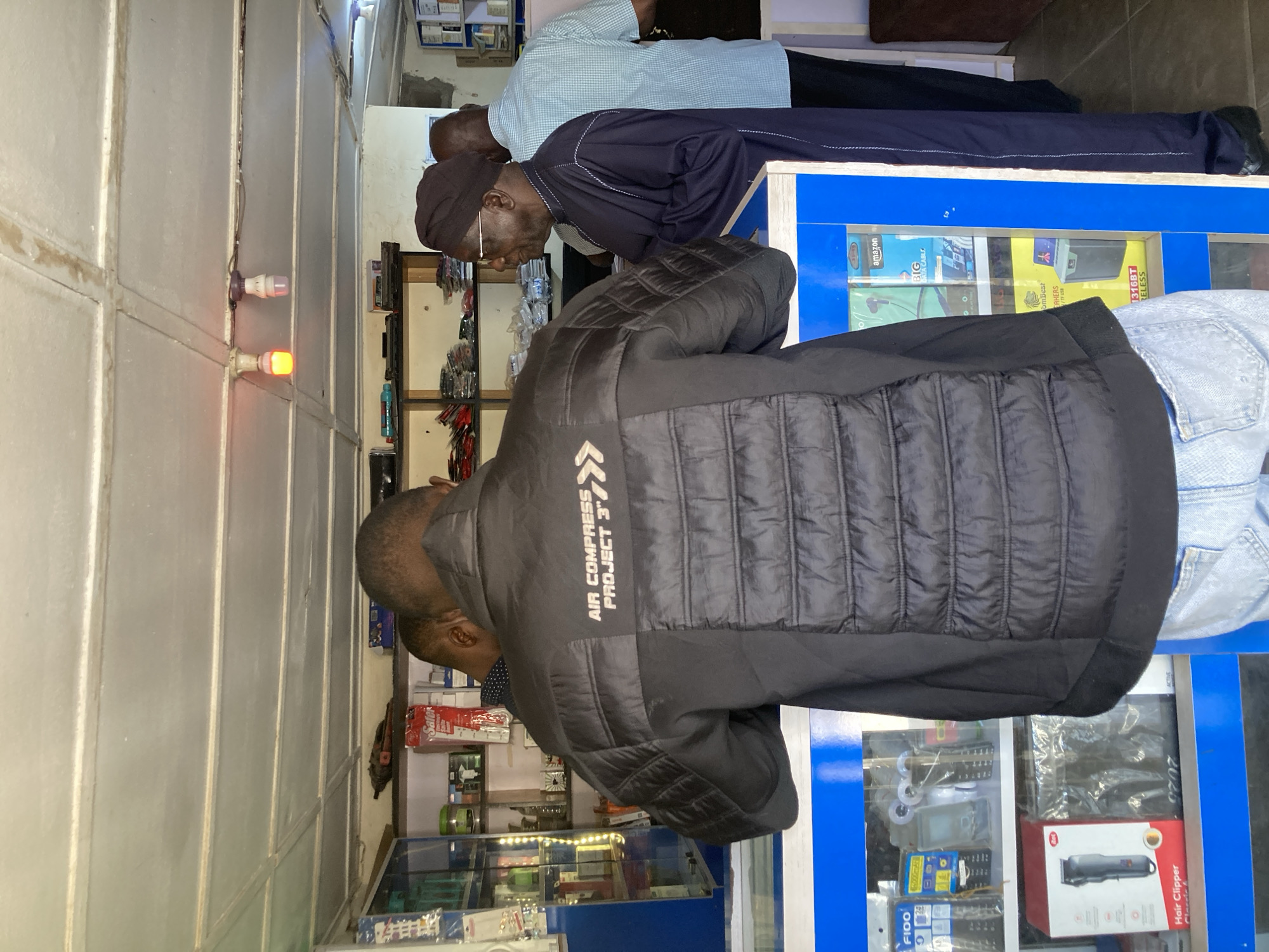 This image captures an indoor scene within what appears to be an electronics or general goods shop in Jos, Nigeria. Three adult men are present, seemingly browsing or shopping.
In the foreground, a man is seen from the back, wearing a black quilted jacket. The jacket features white text on its upper back, reading "AIR COMPRESS PROJECT 3'". He is leaning forward, directing his attention towards a display or counter in front of him.
Behind him, two other men are visible. One man, wearing a dark blue traditional garment and a cap, is also looking down at a blue-framed display counter. Next to him, another man in a light blue checkered shirt stands, similarly observing the products.
The shop's interior features light-colored metal panel walls. A prominent blue-framed display counter holds various packaged items. Text visible on some of these products includes "Hair Clipper," "FIOO," "amazon," "BIG DATA CABLE," and "316BT RELESS" (likely 'wireless'). Other discernible texts include "2020," "Safer," "omBest EAKERS," and "TFUSE." On the wall, two small light bulbs, one glowing red and another white/purple, are mounted. The scene conveys the atmosphere of a local retail establishment.
