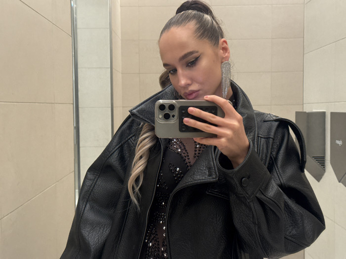 An individual is depicted taking a mirror selfie in what appears to be a public restroom in Dubai, United Arab Emirates. The subject is a female with dark hair pulled back into a high ponytail, revealing large, multi-strand rhinestone drop earrings. She wears a black, oversized leather or faux leather jacket over a dark, sequined, or embellished garment. Her right hand holds a smartphone with a grey textured case, partially obscuring her face, as she poses with slightly pursed lips. The background shows light-colored tiled walls and two grey wall-mounted dispensers, consistent with a commercial or public washroom setting.