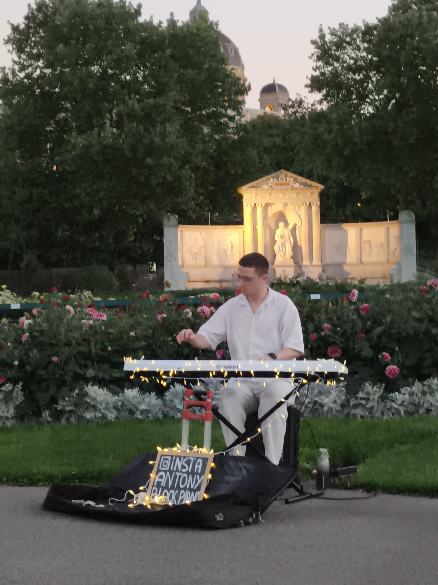 A male pianist wearing glasses, a white short-sleeved shirt, and light-colored pants is seated in front of a digital keyboard adorned with fairy lights in a public park. The keyboard rests on a stand, and a black cloth bag is positioned beneath it. To the left of the bag, a small chalkboard sign with illuminated fairy lights displays "INSTA ANTONY BLOCK PIANO." The background features a lush garden with pink flowers and silver-leafed plants, leading to a stone structure with a statue and ornate architectural details. In the distance, several domed buildings and trees are visible, suggesting an urban setting. The overall lighting indicates it is dusk or early evening. The image is likely taken in the Volksgarten park in Vienna, Austria, given the architectural style of the background structures which resemble elements of the Hofburg Palace complex.