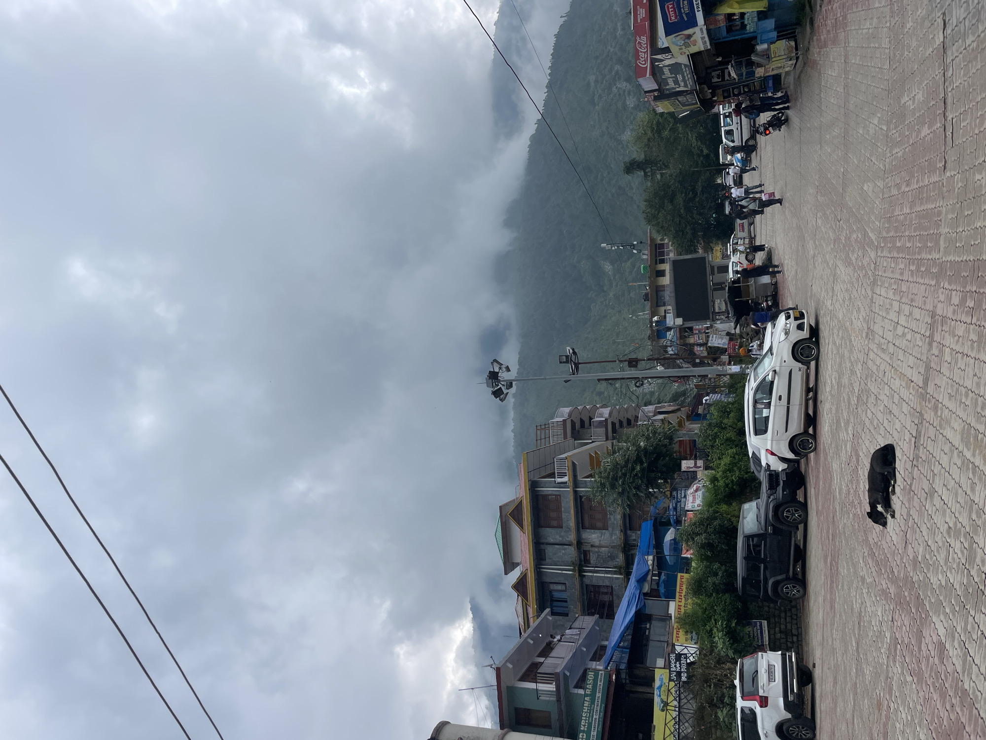 The image depicts a street scene in Bhagsu, India, under a cloudy sky. In the foreground, a black dog lies on a paved area made of interlocking red bricks. To the left of the dog, several cars are parked, including a white SUV and a black Jeep. Beyond the parked cars, a row of multi-story buildings lines the street, with shops and signage visible. One sign reads "KRISHNA RASOI."
Further down the street, more vehicles and people are present. A white van is parked on the right side of the road, next to a Coca-Cola advertisement. Several individuals are walking along the sidewalk, and a street vendor's cart is visible. In the background, lush green mountains are shrouded in thick, gray clouds, suggesting a potentially overcast or rainy day. The architecture of the buildings, with their balconies and colorful facades, is characteristic of the region. The overall atmosphere is that of a bustling, yet somewhat subdued, urban environment at the foothills of the Himalayas.