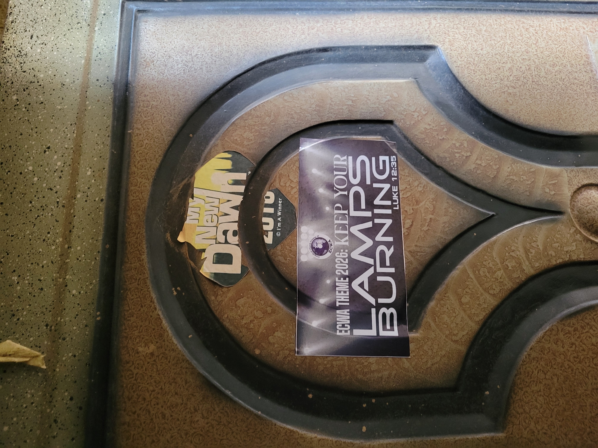 The image shows a close-up of a brown and black ornate surface, possibly a door or a decorative panel. Affixed to this surface are two stickers. One sticker, predominantly yellow, reads "My New Dawn" and "2010 I'm A Winner." The other sticker is black with white text, stating "ECWA THEME 2026: KEEP YOUR LAMPS BURNING LUKE 12:35." The location in the city of Unknown, Unknown cannot be deduced from the image.