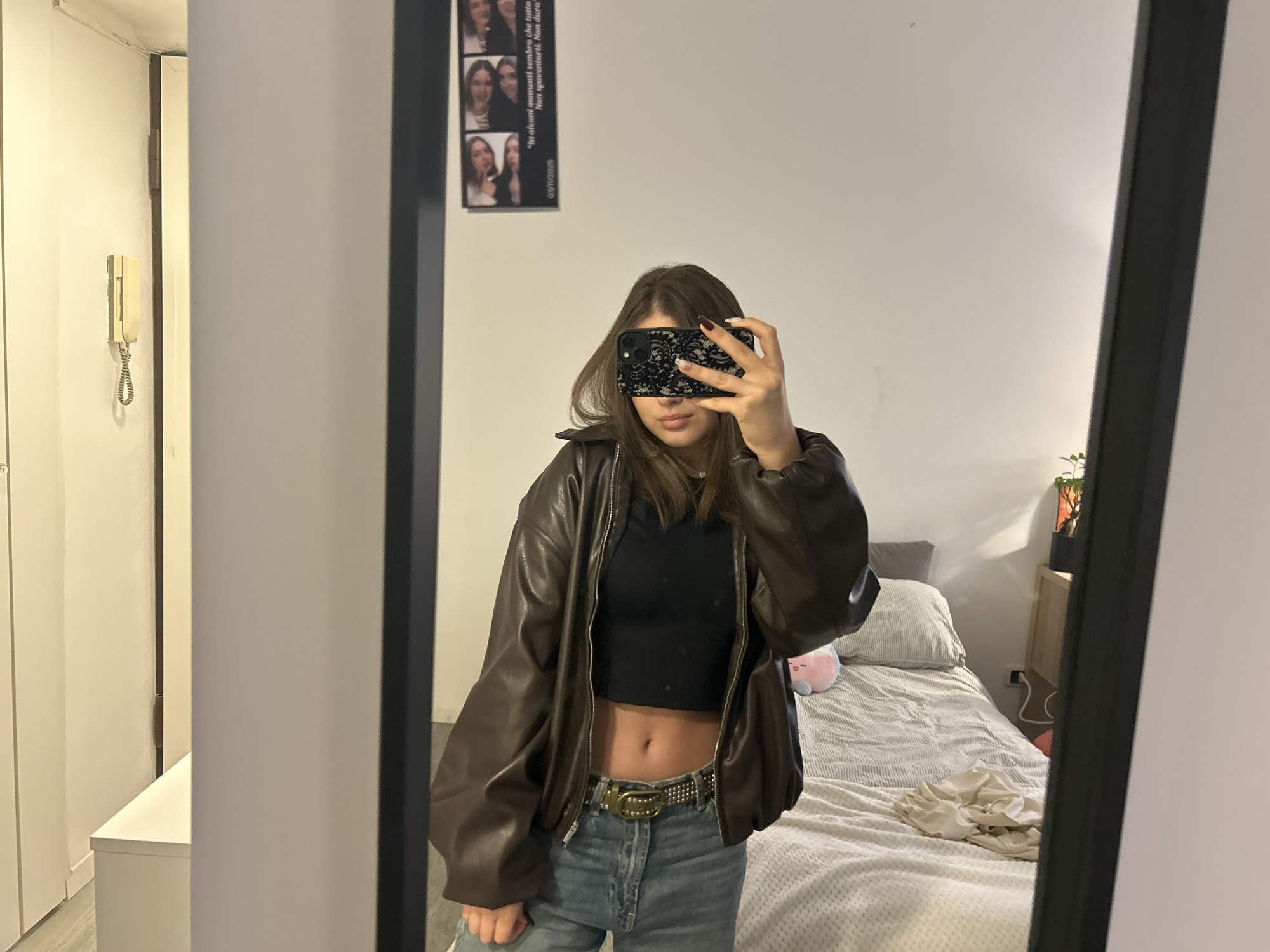 A young woman stands in front of a mirror, holding her phone up to take a selfie. She wears a black crop top, a brown leather jacket, and blue jeans with a studded belt. The background reveals a bedroom with a bed covered in a striped blanket and pillows. A small wooden nightstand with a plant is visible. On the wall to the left of the woman, there is a white door and a small, rectangular poster with images and text. The mirror is framed in black. While the image provides no explicit clues regarding the location, there's no way to confirm it is in Milan, Italy without additional information.