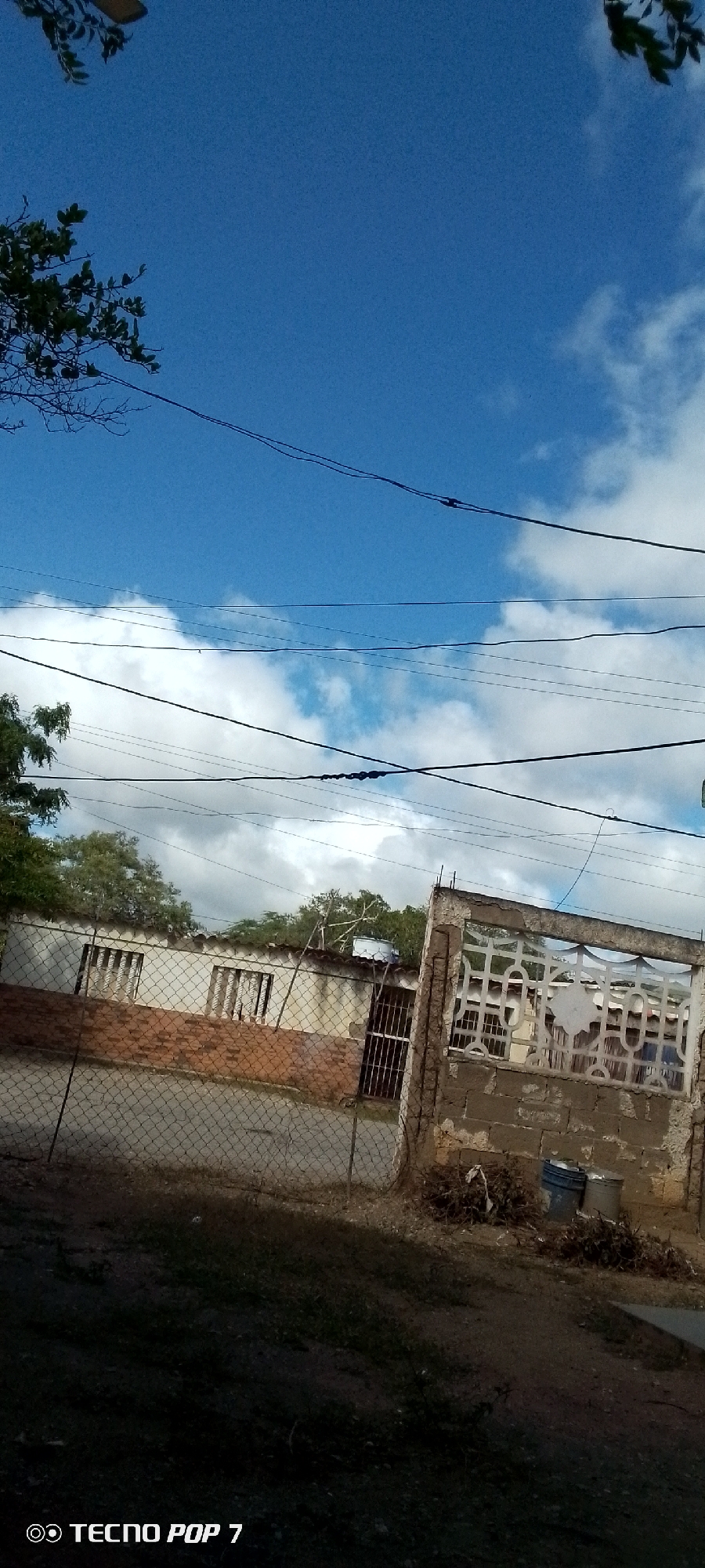This is an outdoor photograph taken during the daytime in Altagracia, Venezuela. The scene depicts a street view with a building in the background, partially obscured by a wire fence in the foreground. The building appears to be a modest, single-story structure with a white facade and a red-tiled roof. It has barred windows and a section of exposed brickwork. To the right of the building is a partial view of a concrete structure with an ornamental railing. In front of this structure, there are two plastic buckets and a pile of dried brush. The ground in the foreground is covered with vegetation. Above the building, numerous power lines crisscross the frame against a bright blue sky dotted with white clouds. The image is captured from a slightly elevated perspective looking downwards.