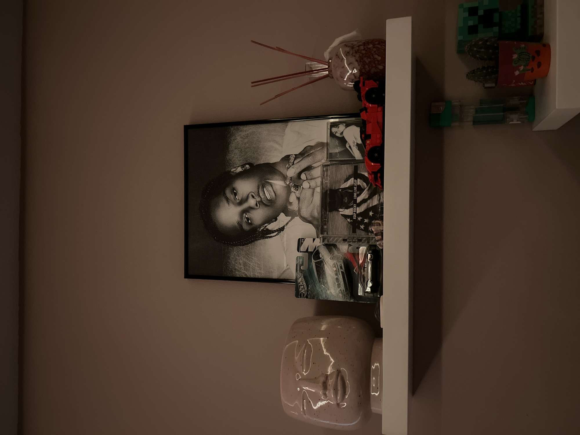 The image shows a wall shelf in a room. On the shelf are several objects, including a framed black and white photograph of A$AP Rocky, a reed diffuser, a red toy car, two toy cacti, a toy Minecraft Creeper, and a ceramic vase shaped like a human face. The wall is painted a light brown color. The photo appears to have a few other CDs or albums included in it as well, and two hot wheels cars. There is no information in the image that suggests the location is in Goce Delchev, Bulgaria.